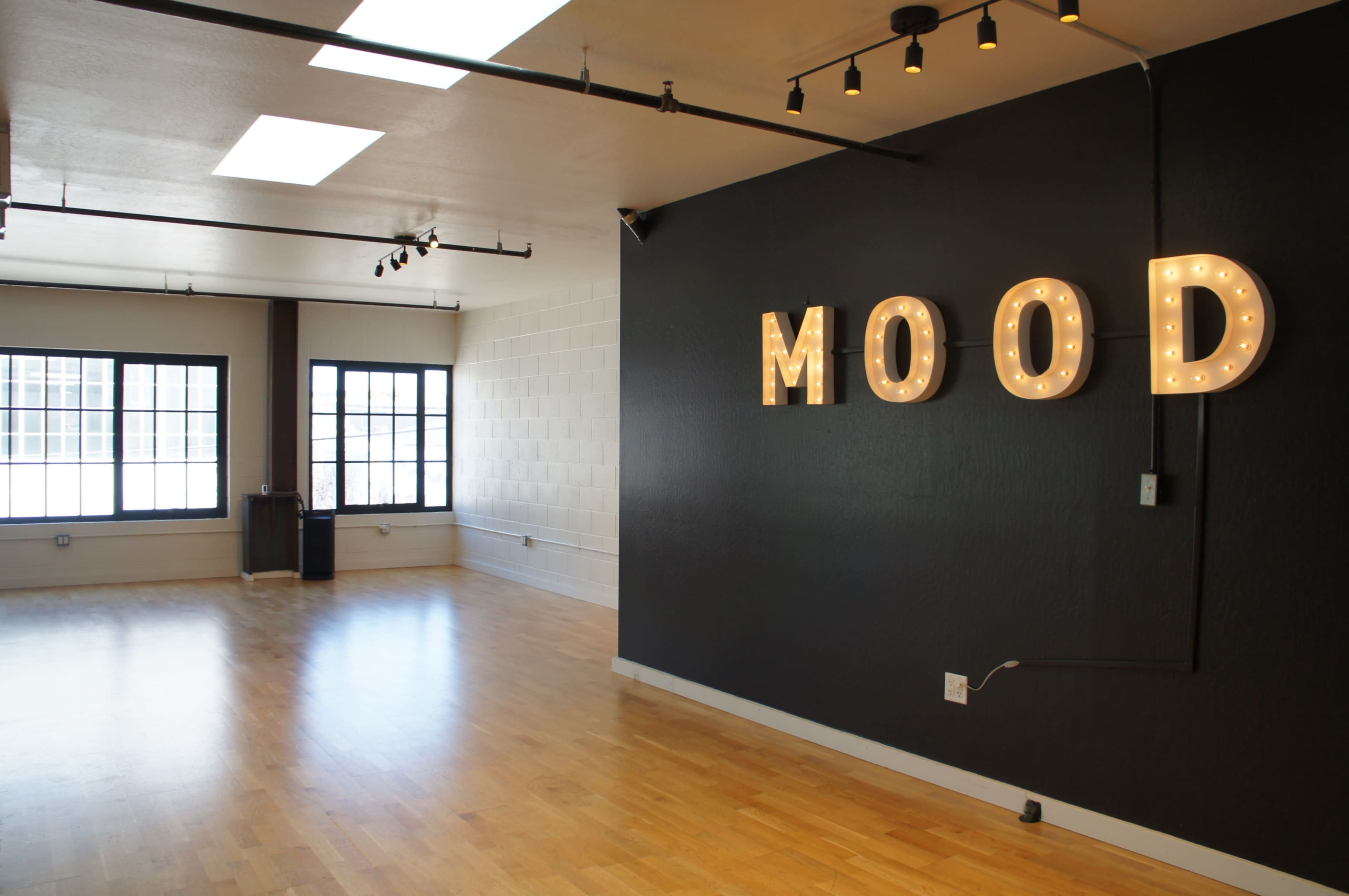 The image shows a spacious, well-lit room featuring a black wall with illuminated letters spelling "MOOD" and large windows that let in natural light.
