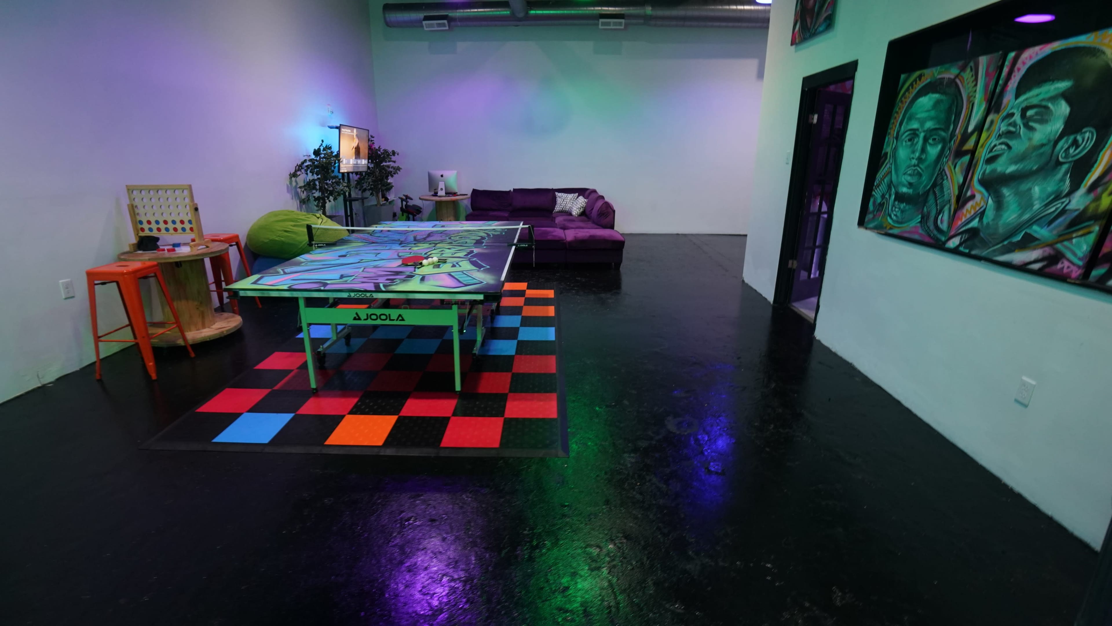 A game room featuring a colorful ping pong table in the center, surrounded by a purple sofa and vibrant artwork on the walls.