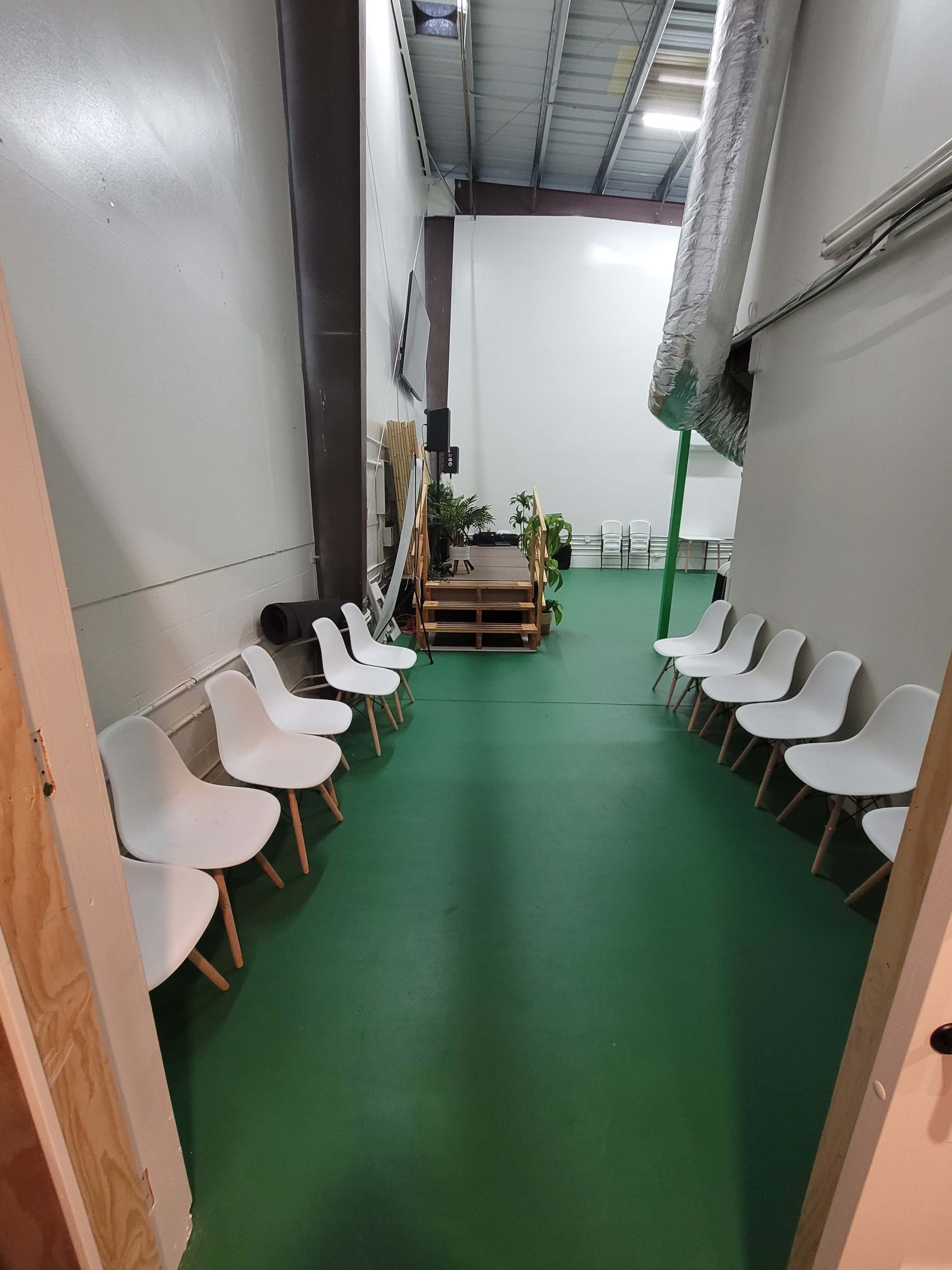 The image shows a narrow hallway leading to a spacious room with several white chairs aligned on the left and a stack of wooden pallets at the end.
