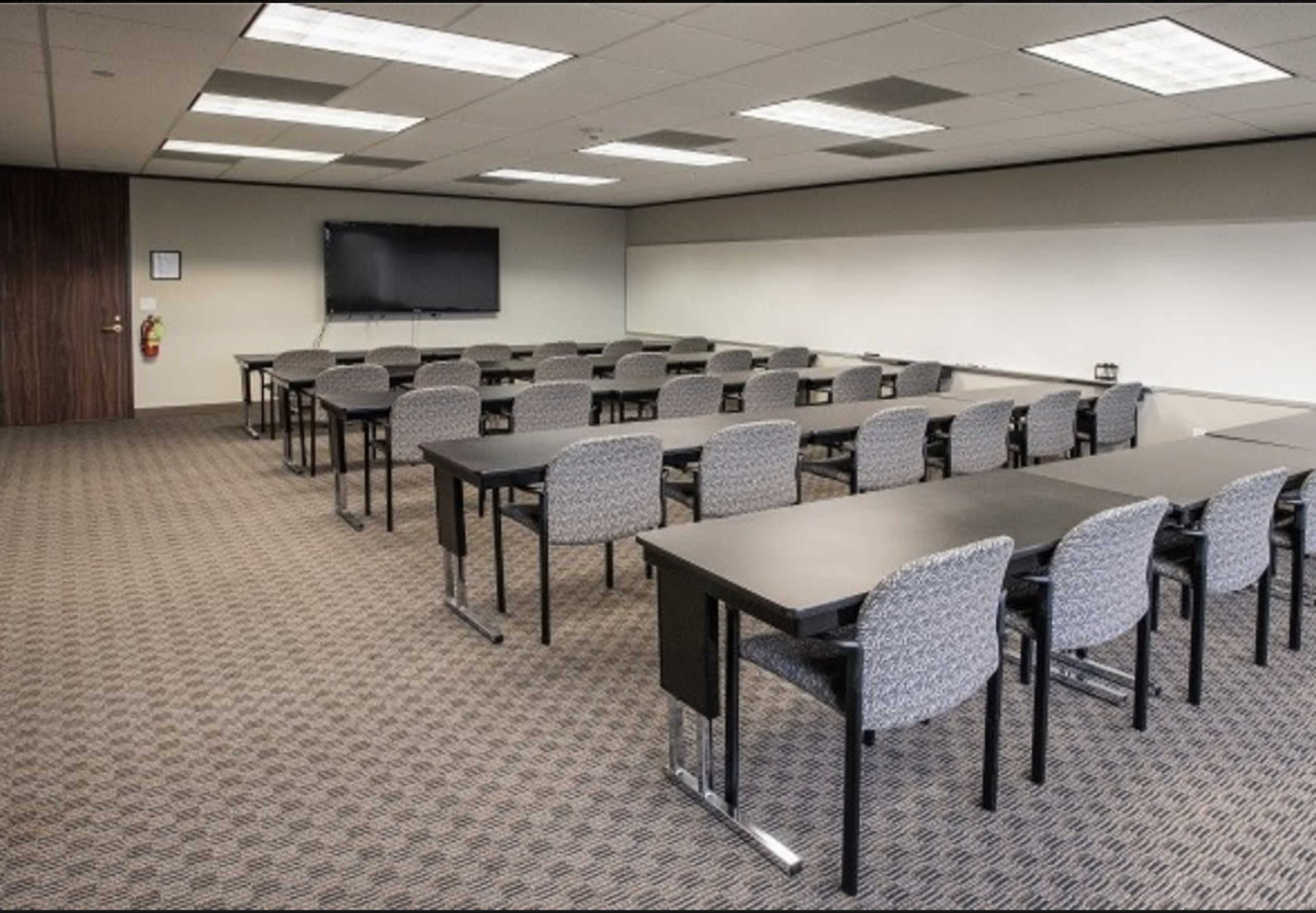 A modern conference room features rows of black tables and grey chairs with a large monitor mounted on the wall.