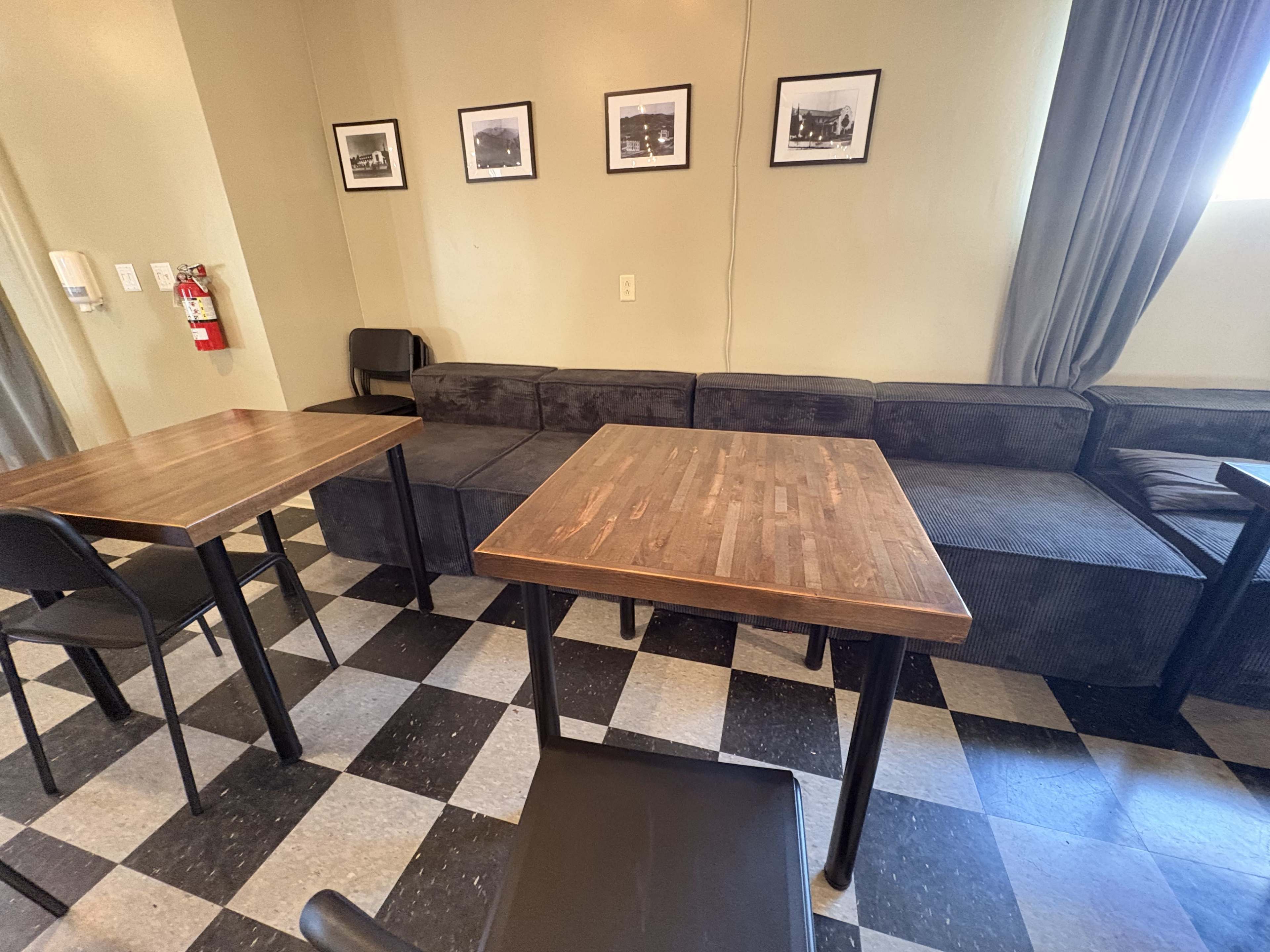 The image shows a cozy cafe interior featuring two wooden tables, black chairs, and a cushioned seating area against the wall, with framed photographs hung above.