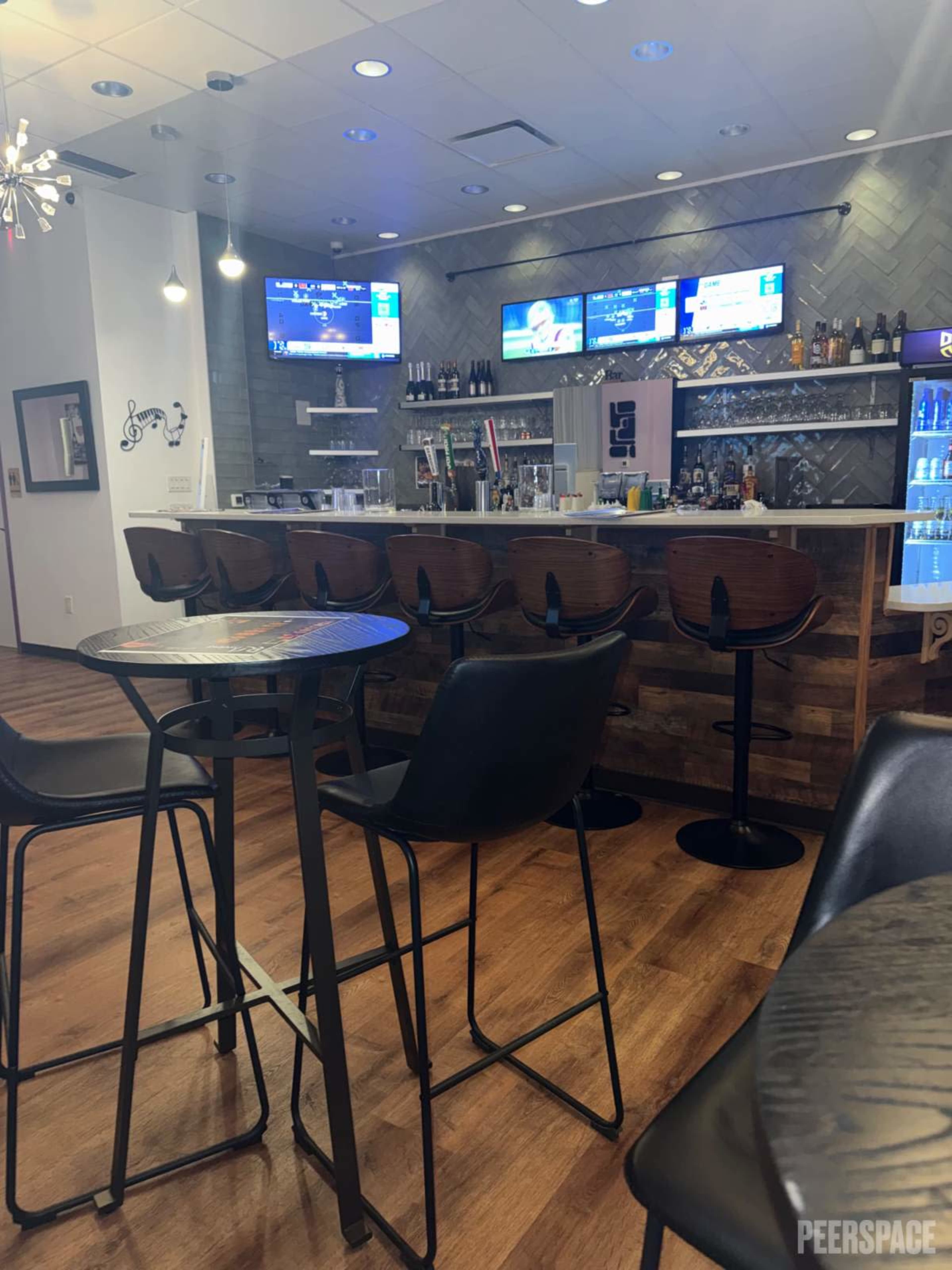 The image shows a modern bar area with high tables and stools, a polished counter, and several screens displaying information.