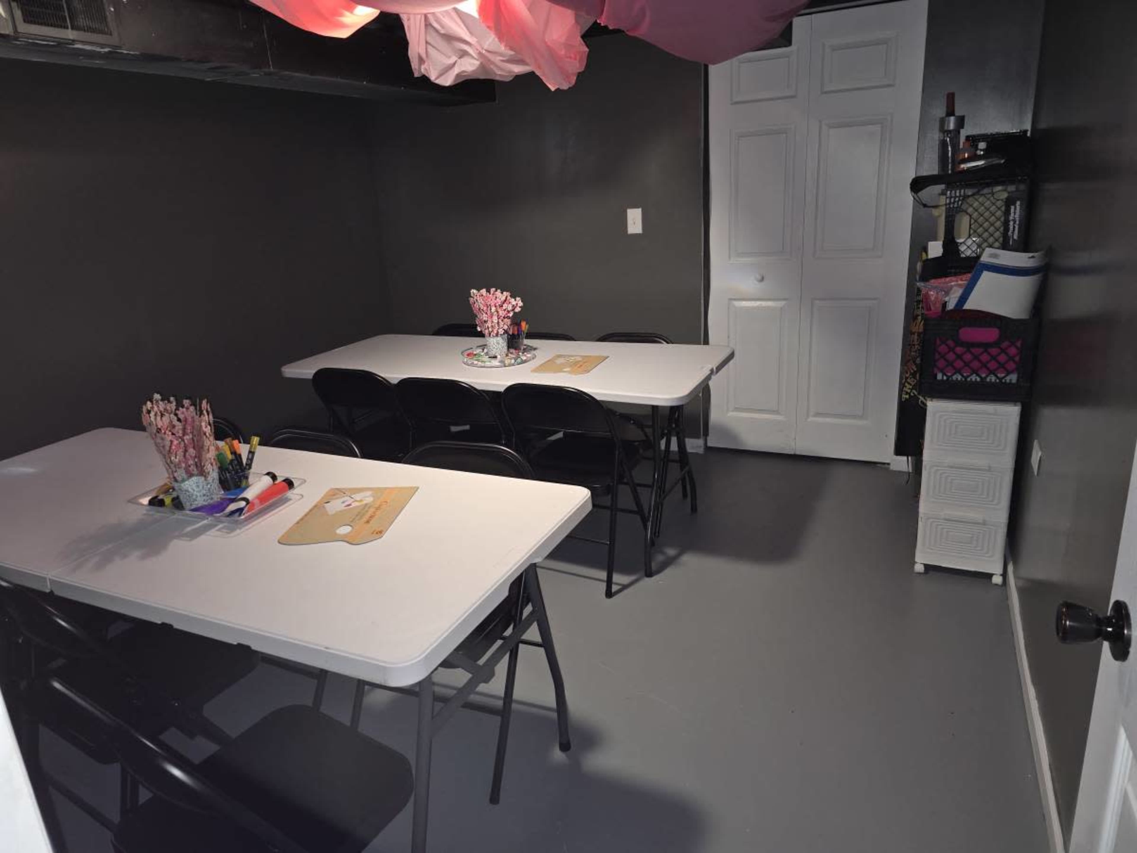 The image shows a small room with two tables and black folding chairs arranged neatly, along with various items on the tables and shelves.