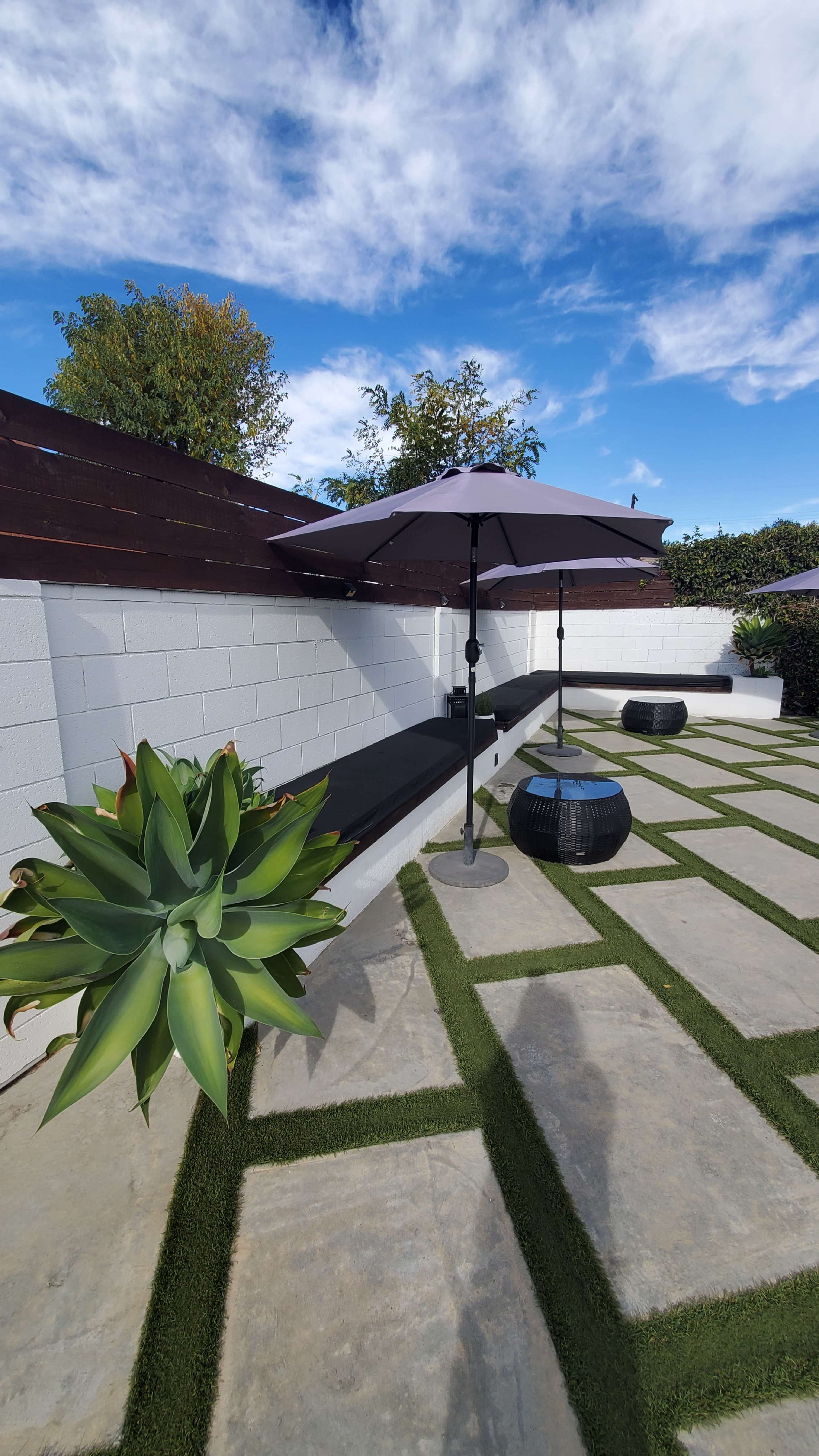 The image shows a landscaped outdoor area with a stone patio, two shaded umbrellas, and a large succulent plant in the foreground.