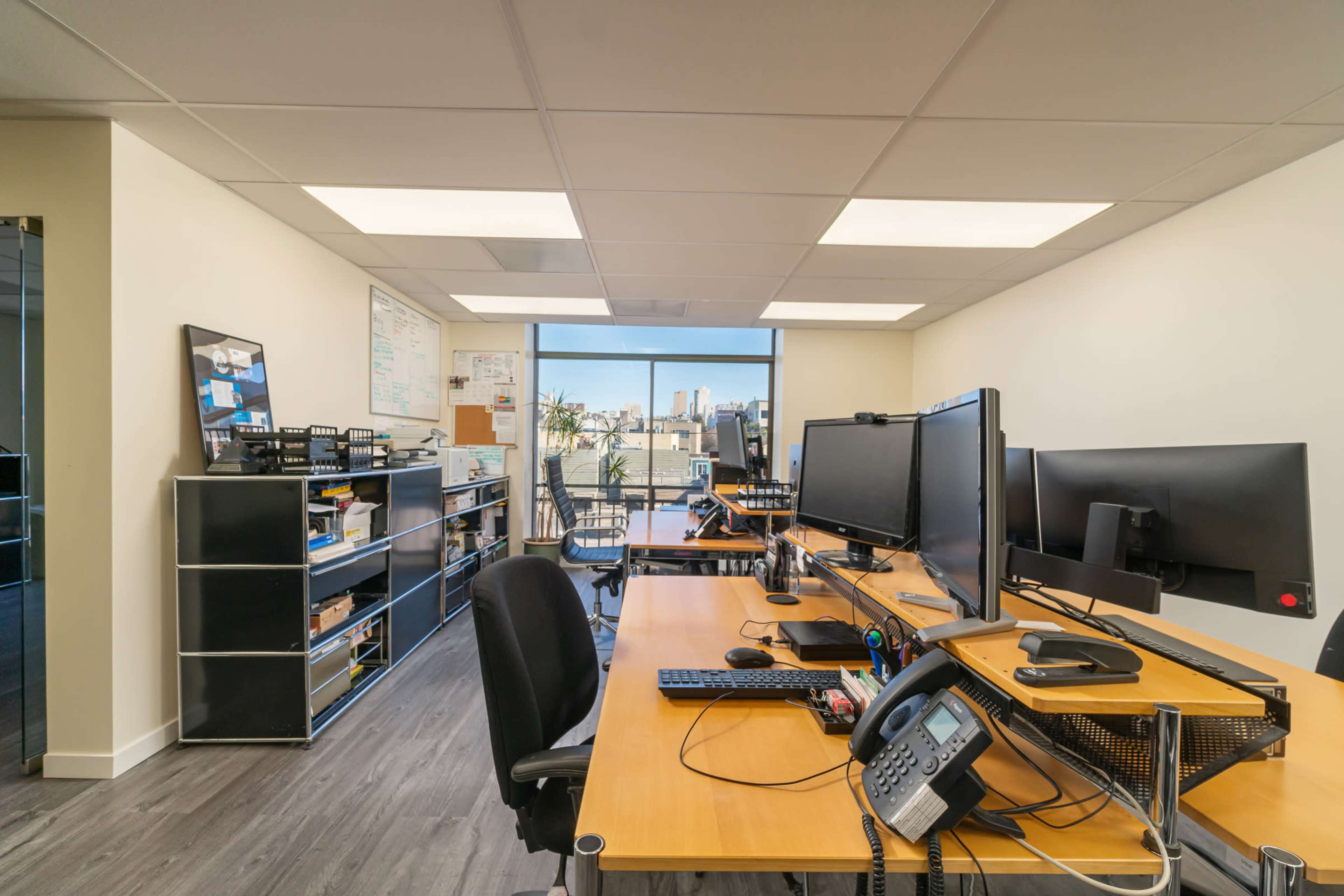 A modern office space with two large computer monitors on a wooden desk, shelves filled with office supplies, and a view of the city skyline through a large window.