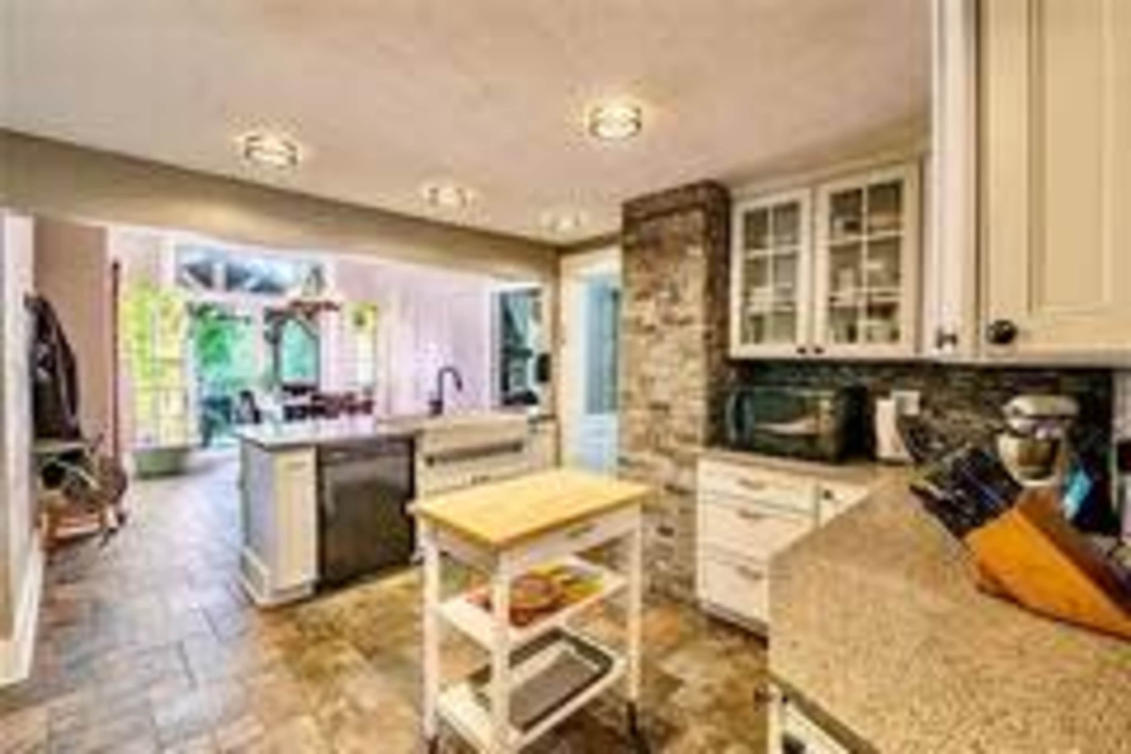 The image shows a modern kitchen with an island, granite countertops, and a combination of stone and white cabinetry, featuring natural light flowing in from large windows.