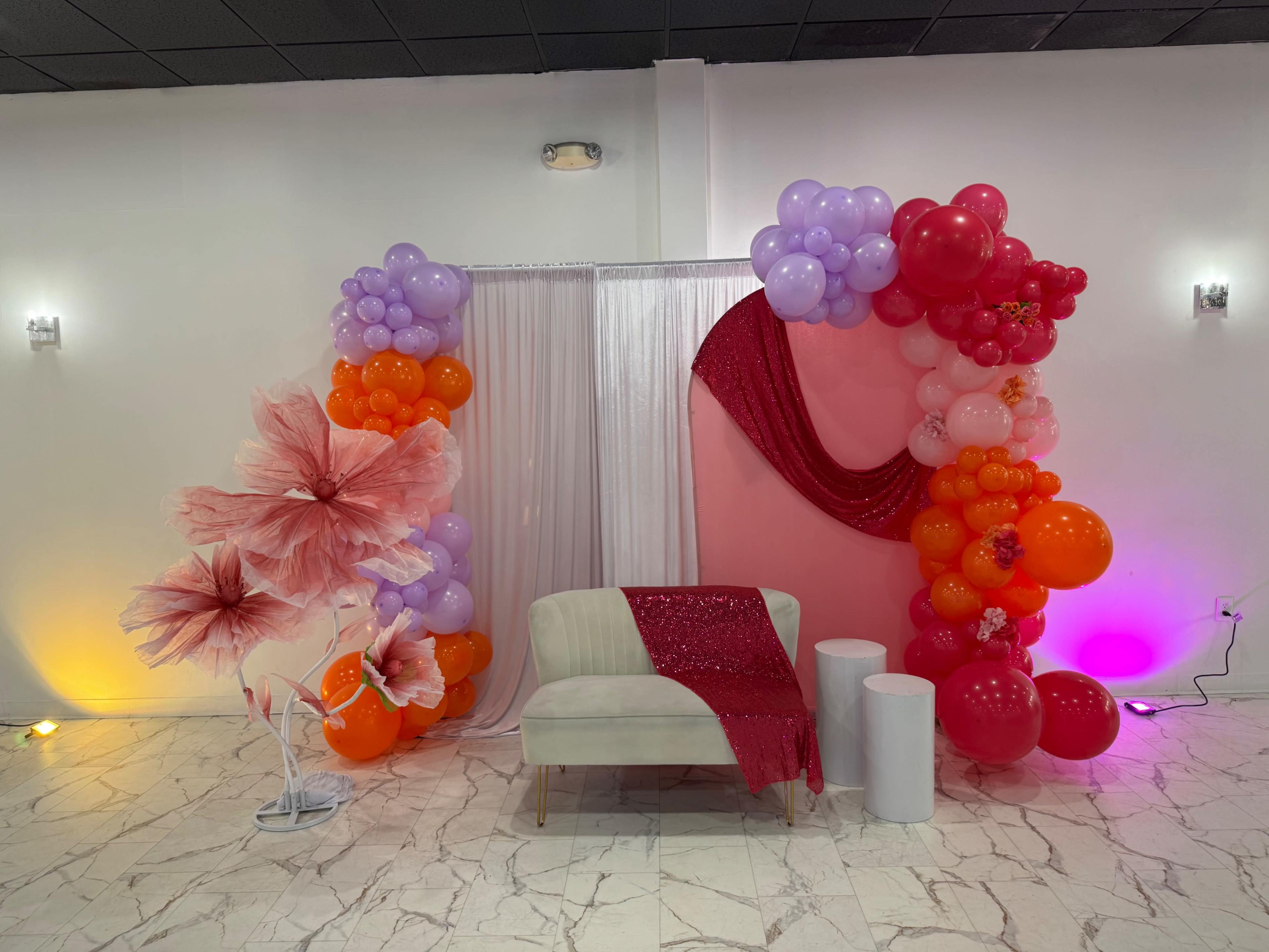 The image depicts a decorated event space featuring a backdrop of balloons in various colors, a white curtain, a sparkly red drape, large floral displays, and a cream-colored sofa.