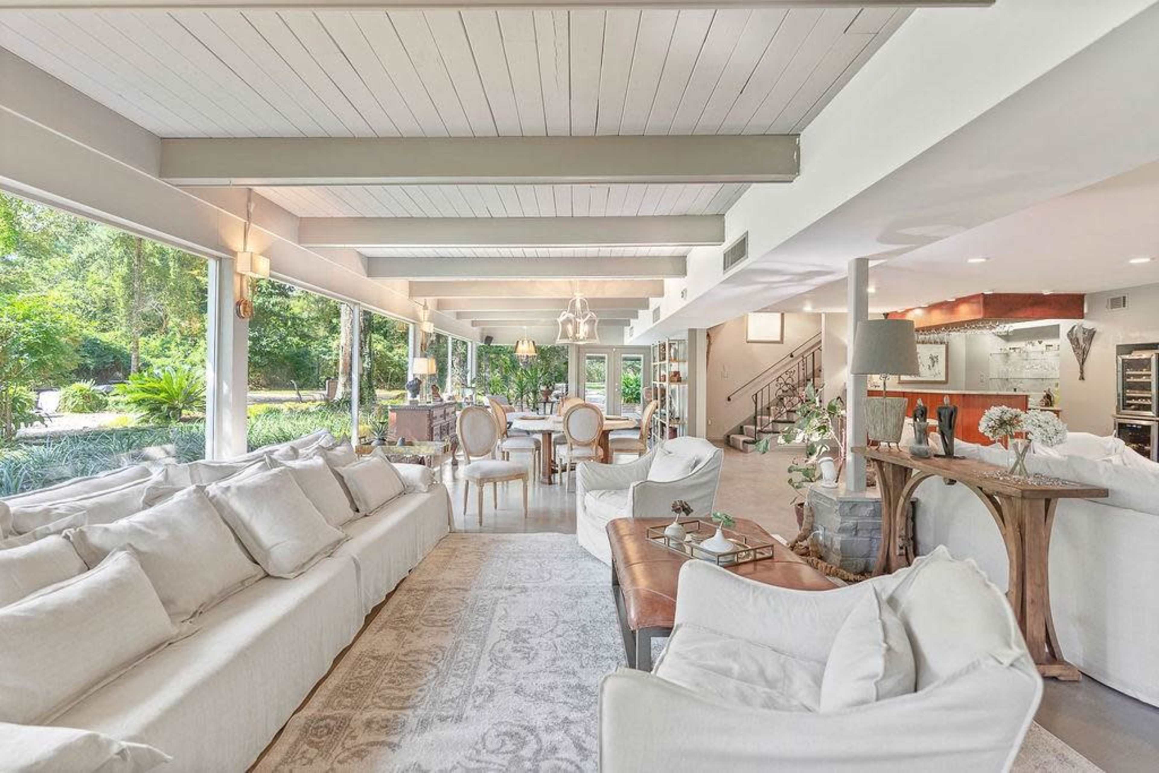 A spacious living area features large windows, light-colored furniture, and an open layout with a dining area and visible kitchen.