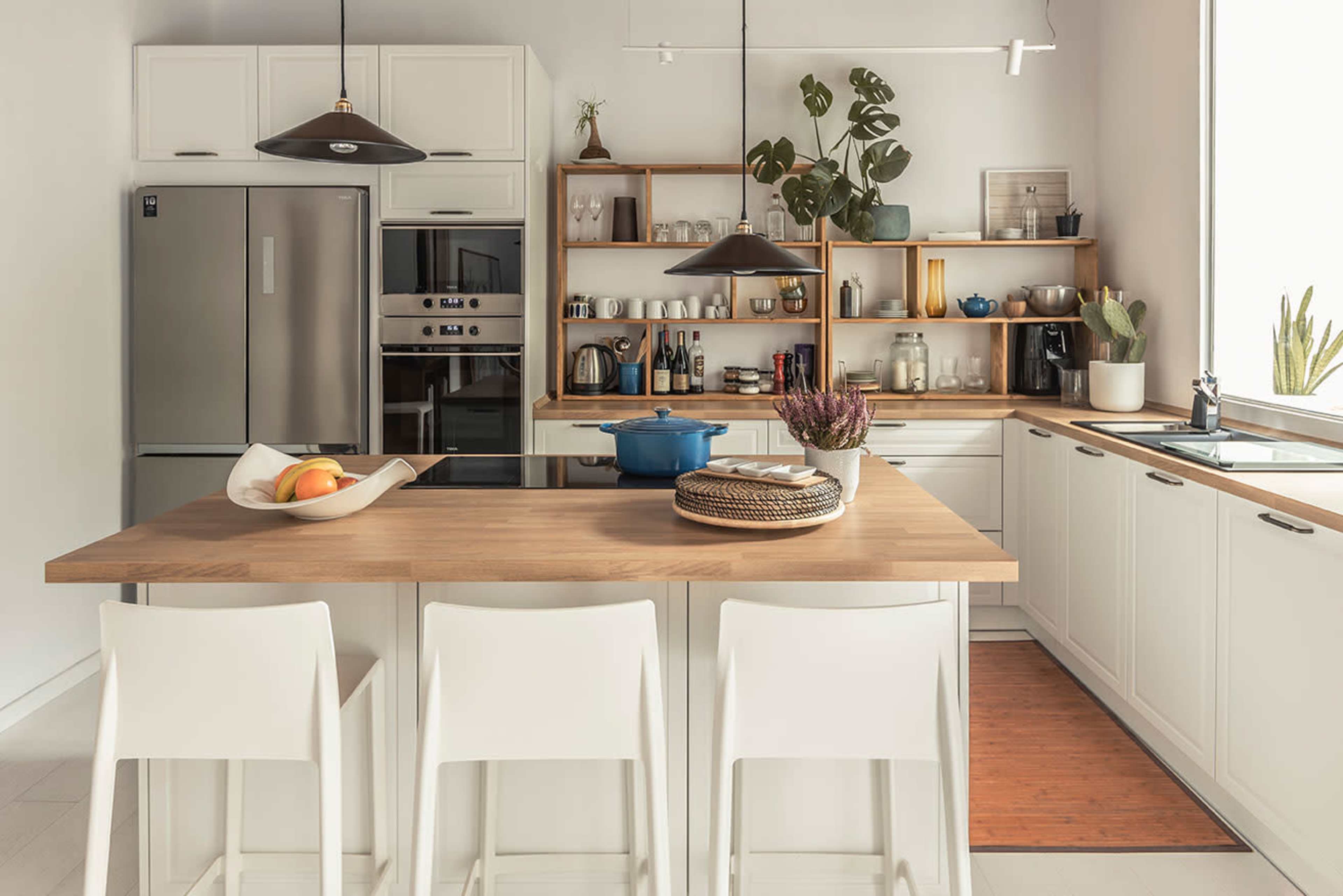 A modern kitchen with a central wooden island, three white bar stools, stainless steel appliances, and a shelf displaying various kitchen items and plants.