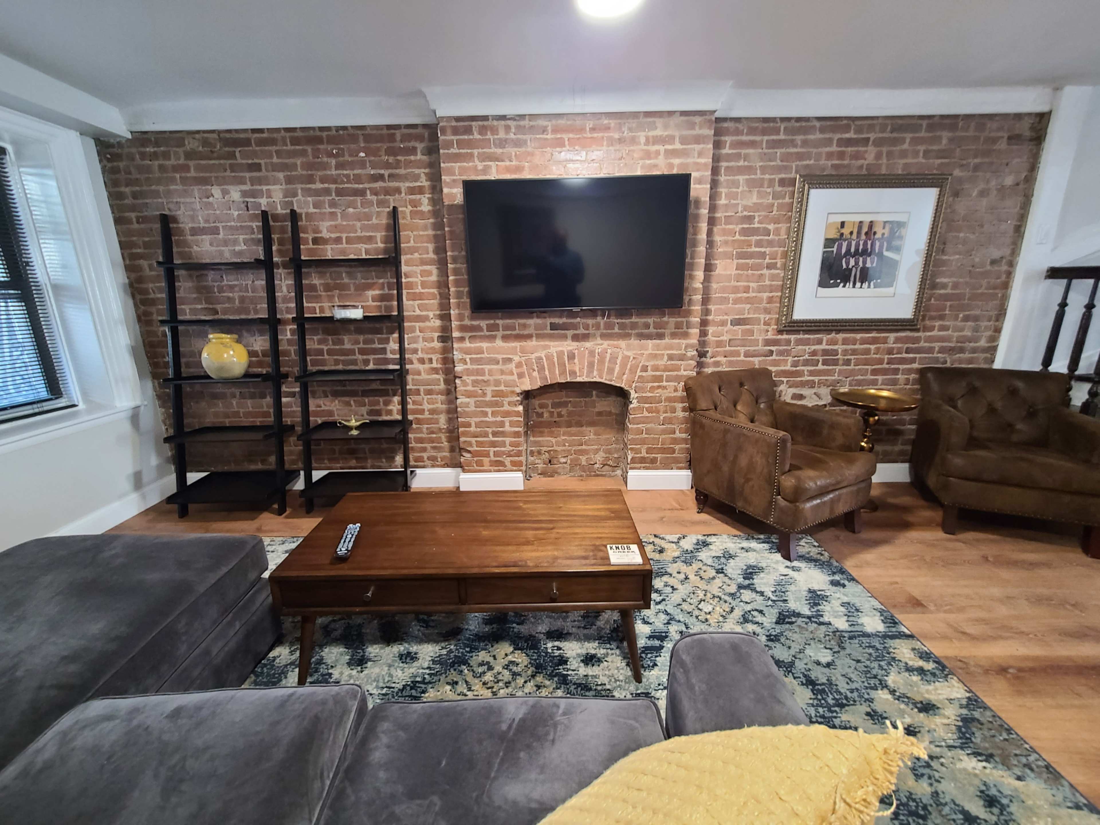 The image shows a living room with exposed brick walls, a television mounted above a brick fireplace, and a coffee table surrounded by brown leather chairs and a yellow throw blanket.