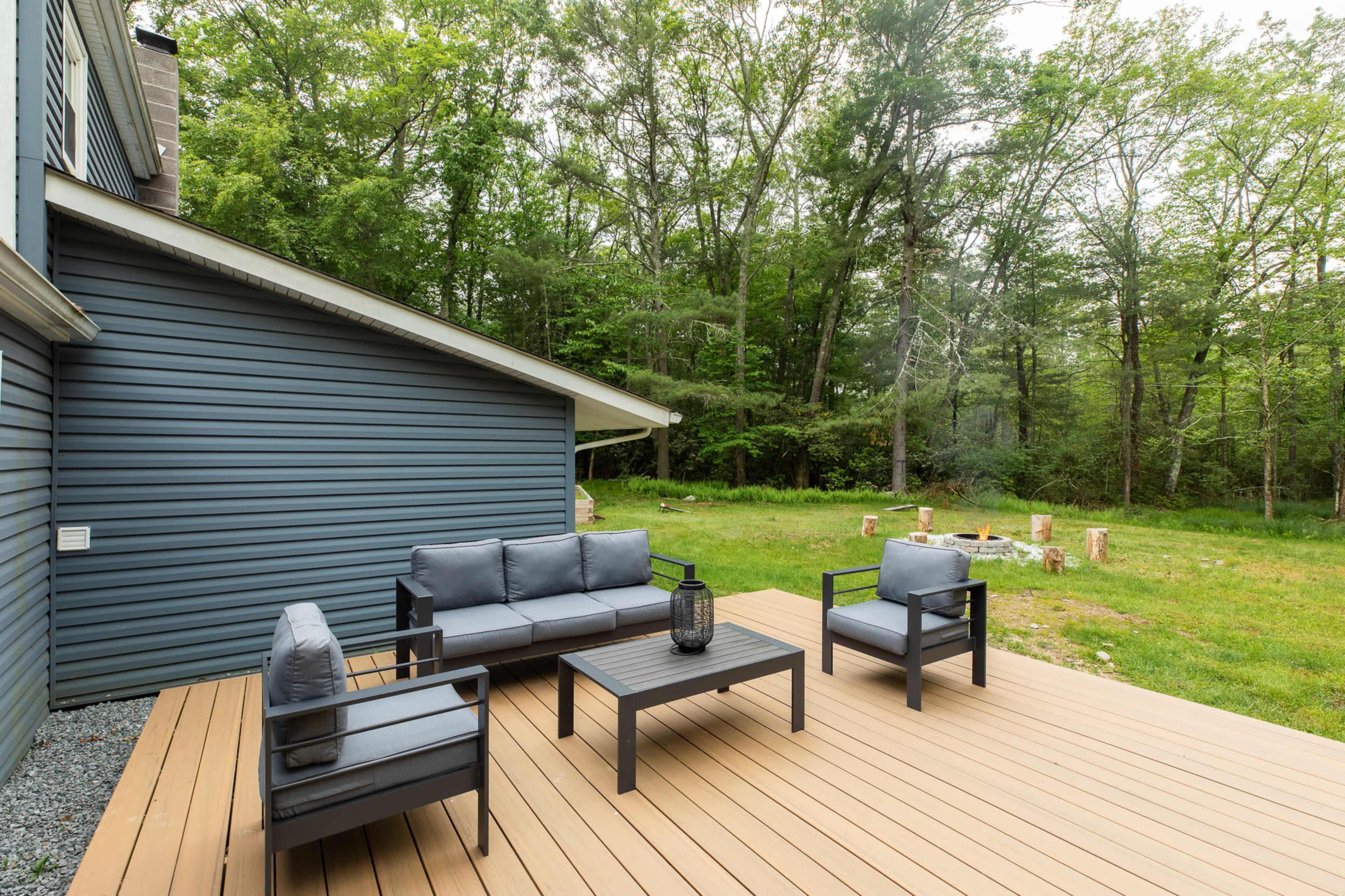A modern outdoor seating area features a black couch and chairs around a wooden coffee table on a deck adjacent to a grassy area and trees.