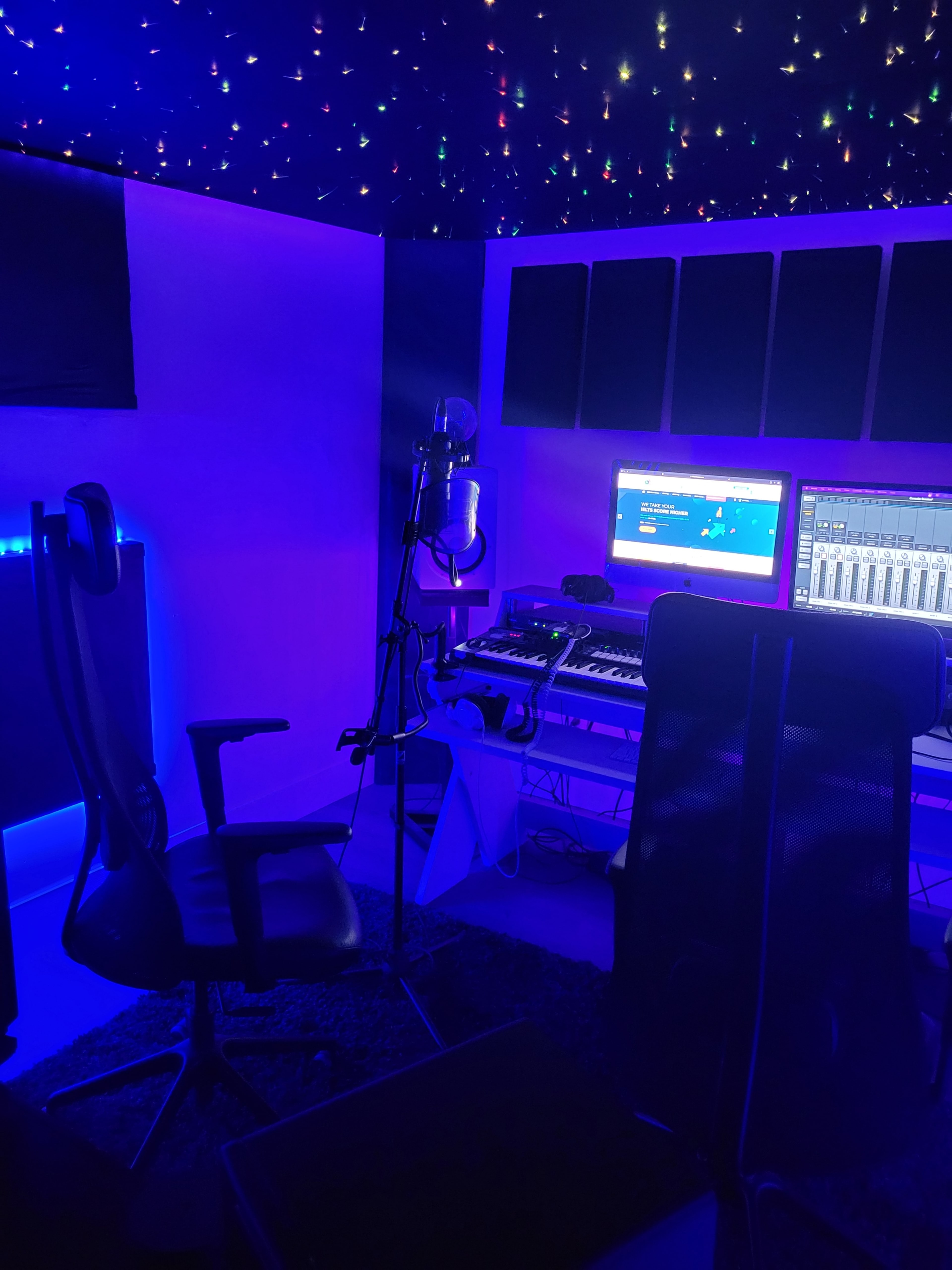 The image shows a dimly lit music studio featuring a microphone, keyboard, computer monitors, and a chair, all surrounded by a starry ceiling.