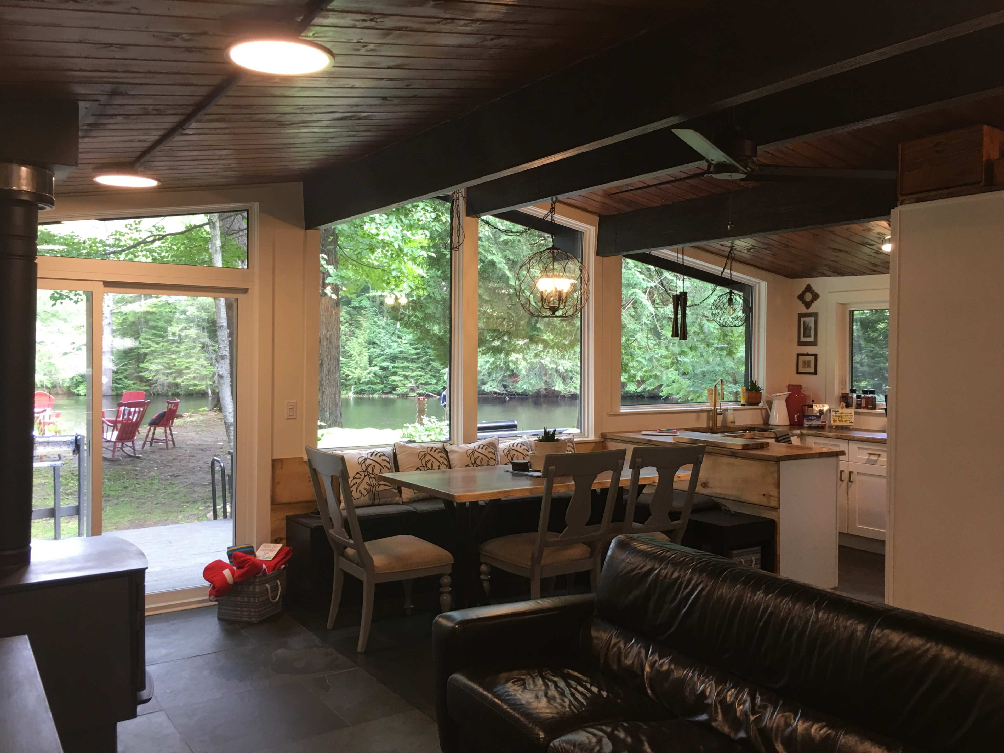 A bright, open kitchen area with a dining table, overlooking a lake and wooded landscape through large windows.