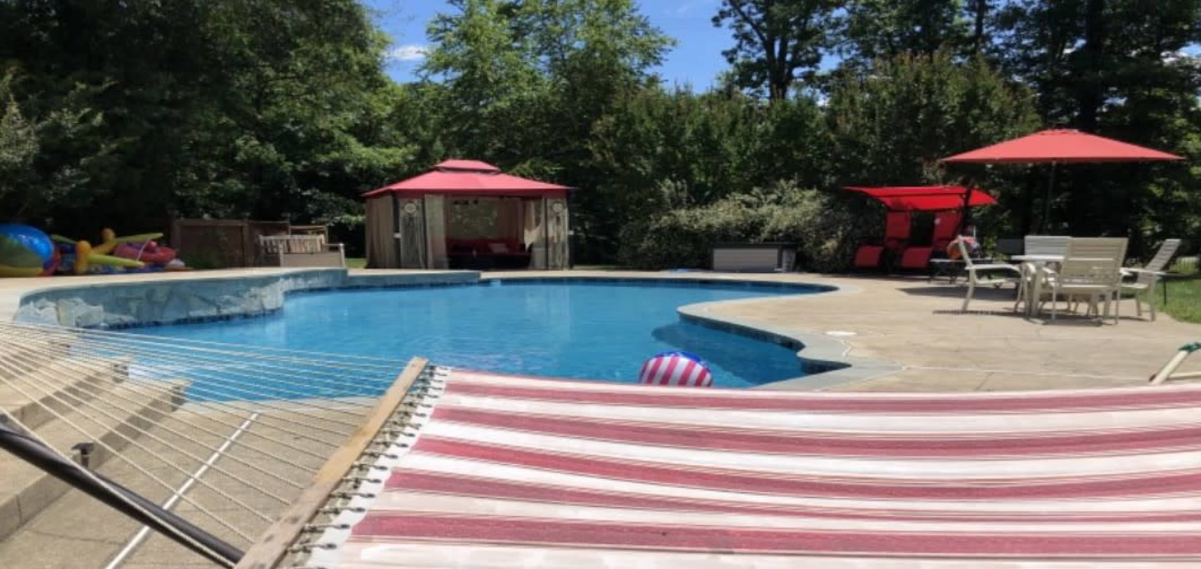 A backyard pool area with a blue swimming pool, two red canopies, lounge chairs, and a hammock in the foreground.