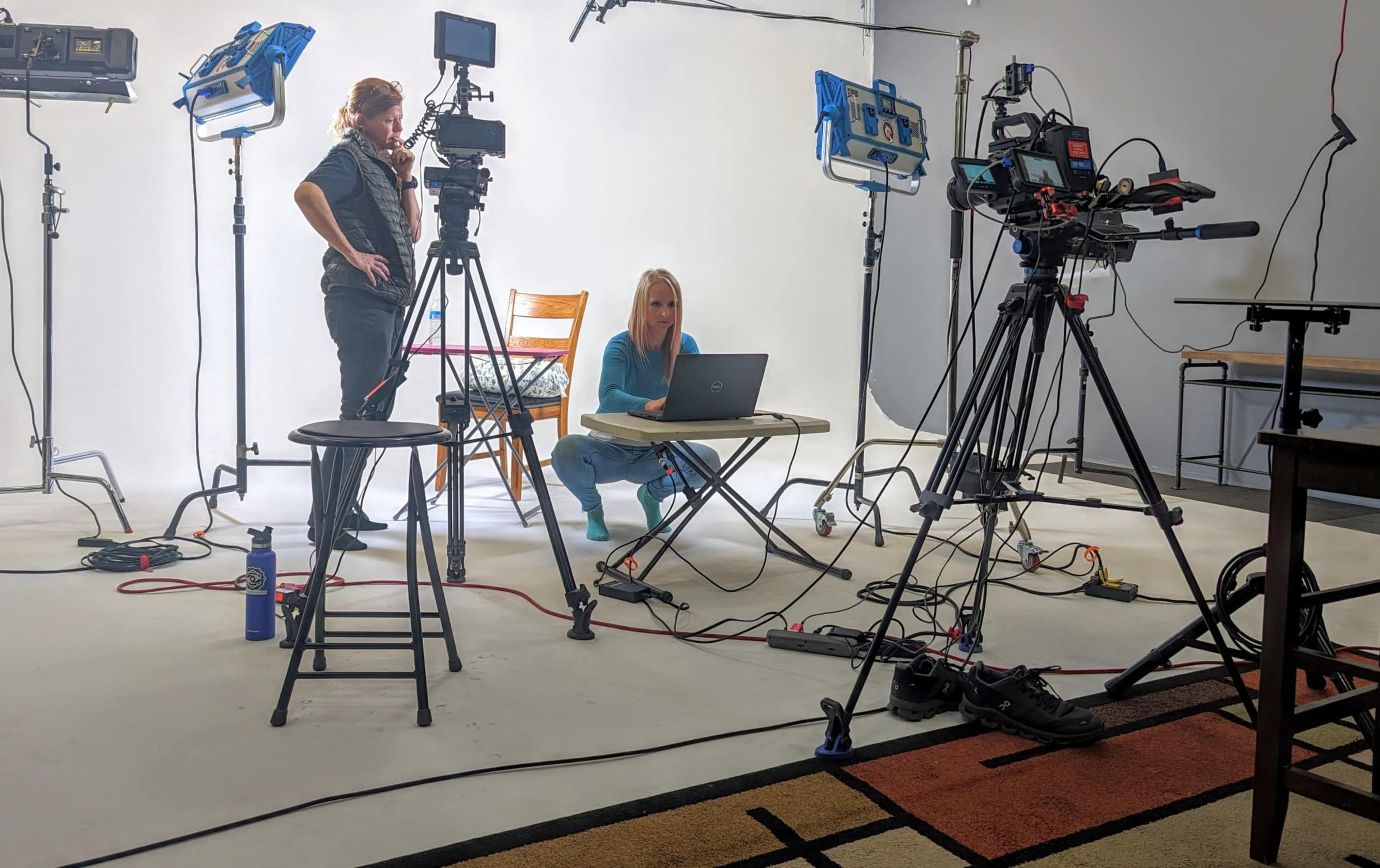 A woman sits at a table with a laptop while a man stands nearby, surrounded by multiple cameras and studio lights.
