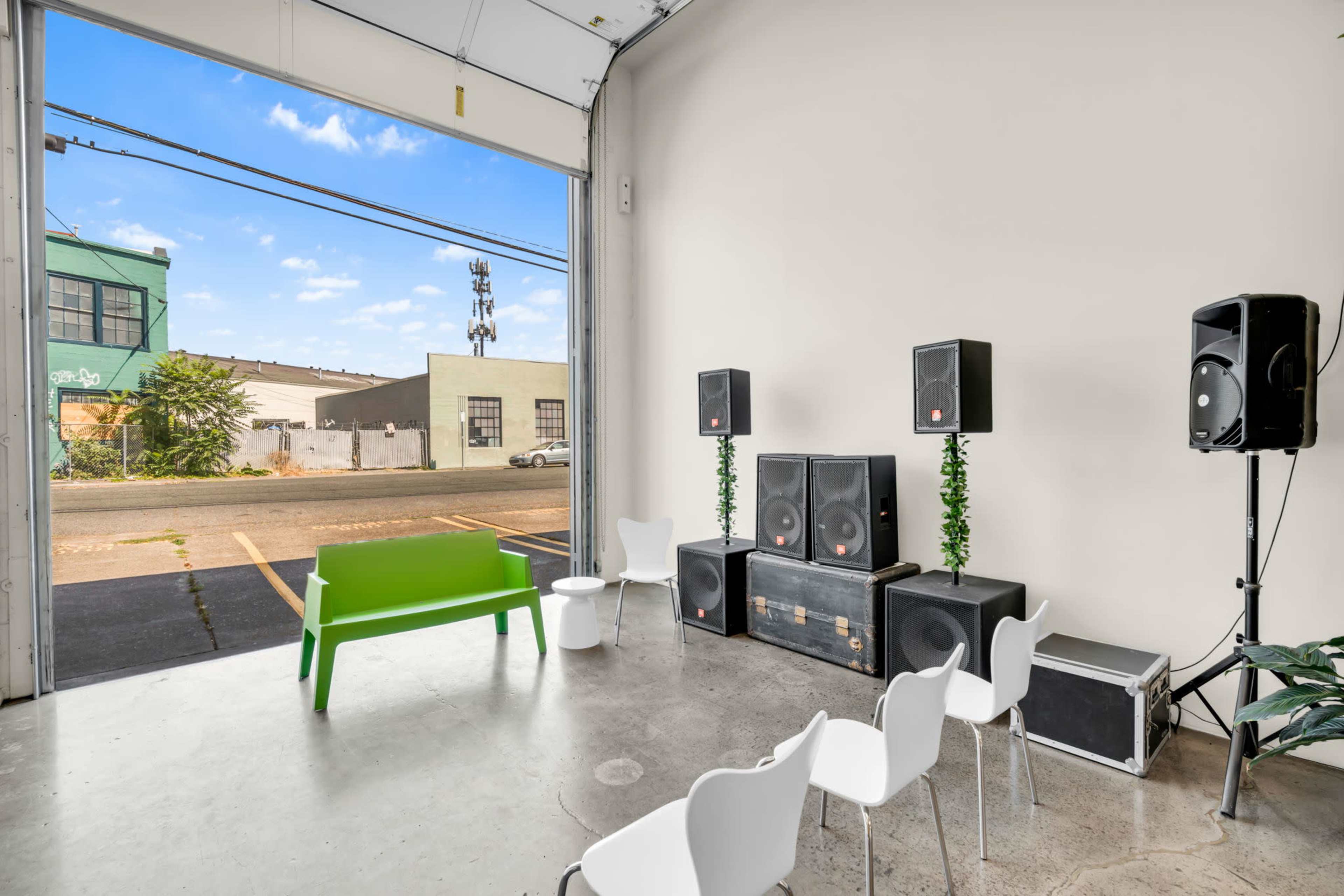 The interior of a warehouse features a set of white chairs arranged in front of large speakers and a green bench, with a view of an outdoor street scene through an open door.