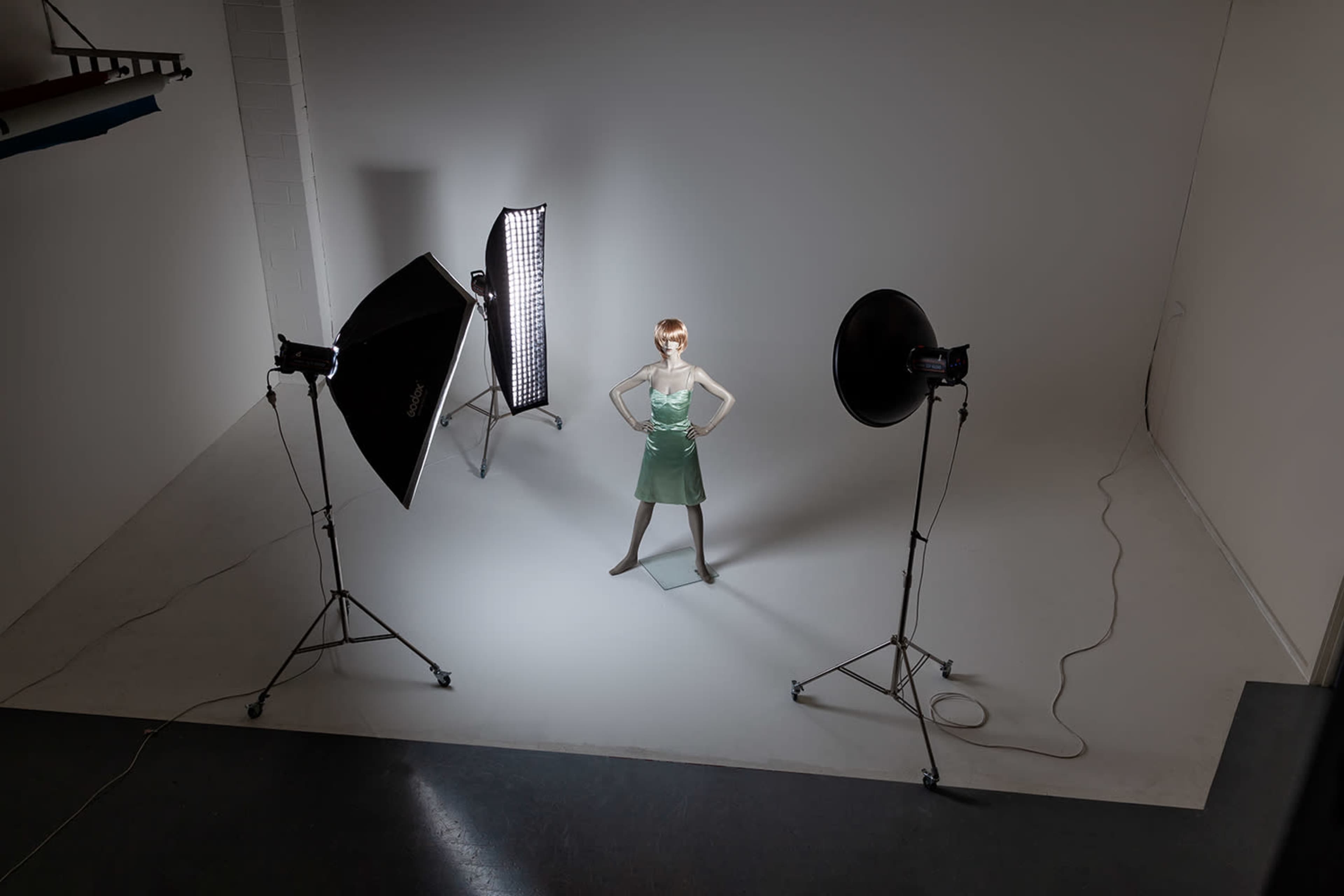 A mannequin dressed in a green dress stands in the center of a photography studio surrounded by lighting equipment.
