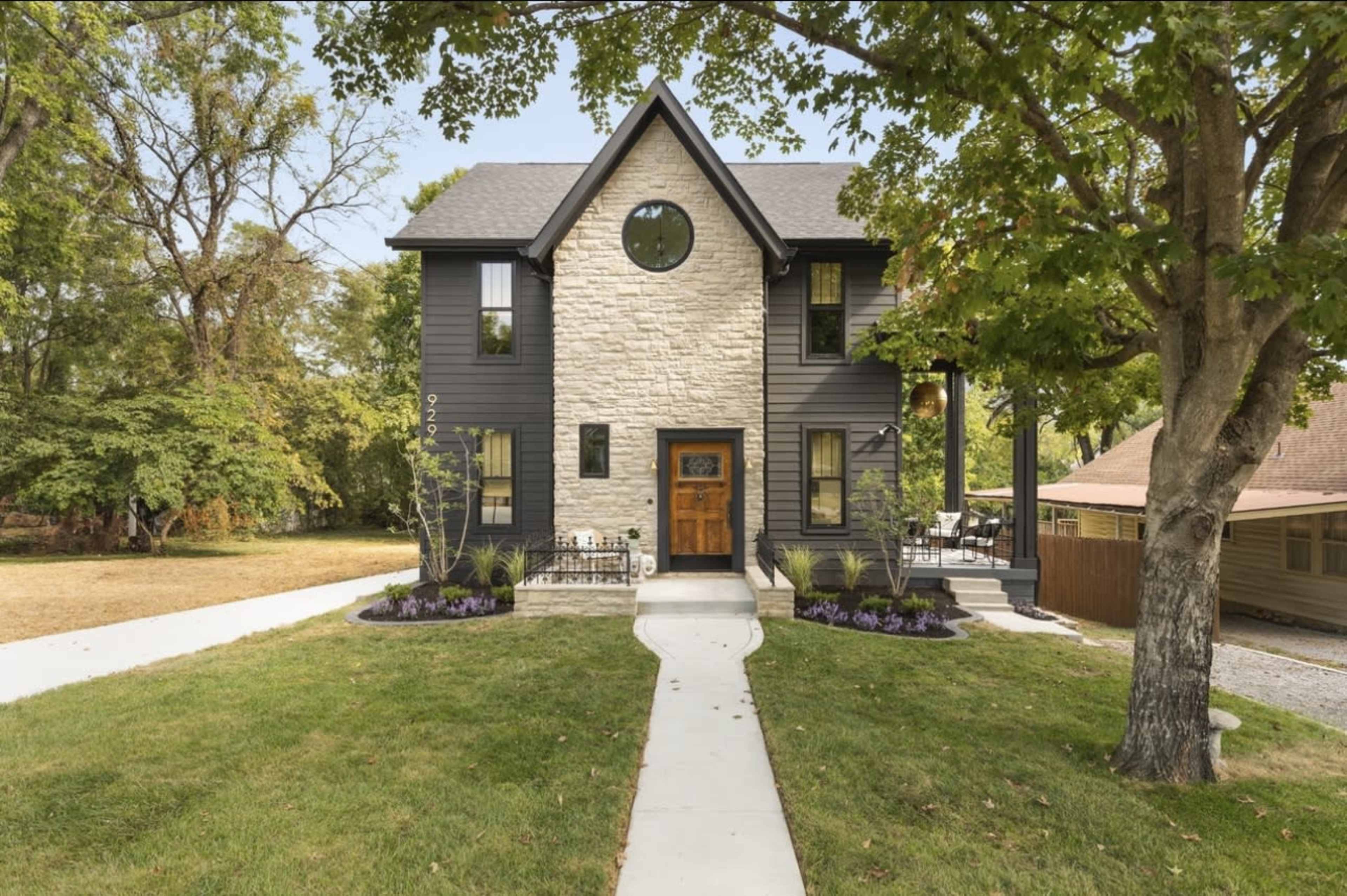 A two-story stone and wood exterior house with a circular window, surrounded by landscaped greenery and a concrete pathway.