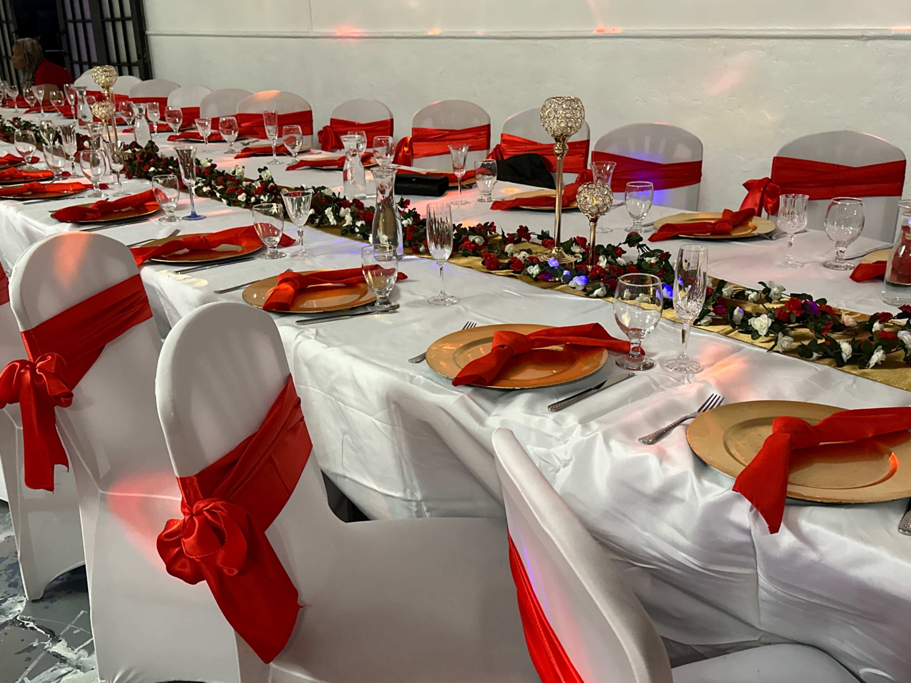 A long banquet table is decorated with white tablecloths, red chair sashes, and gold plates, surrounded by glasses and floral centerpieces.