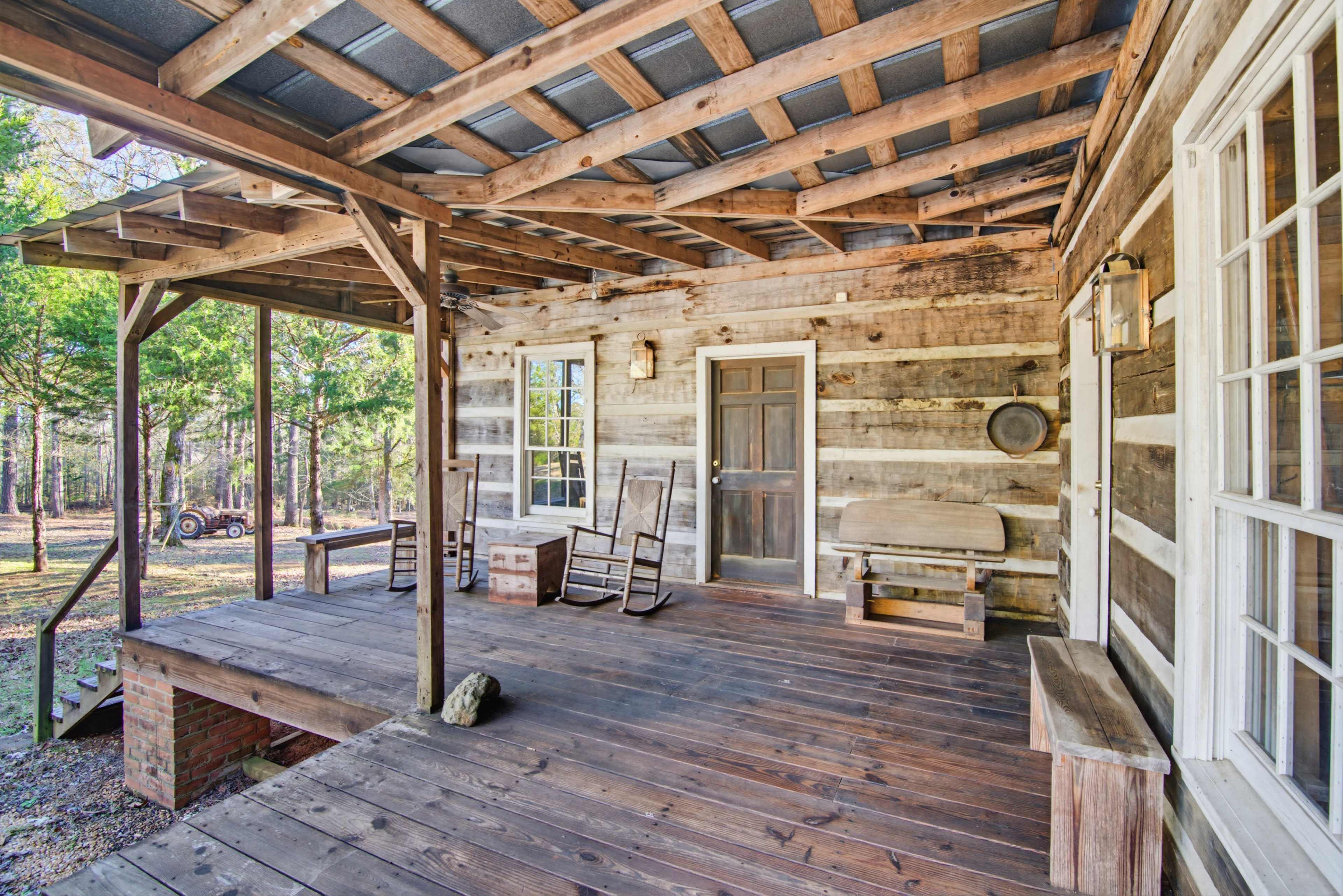 The image shows a wooden porch with several rocking chairs and a bench, attached to a log cabin surrounded by trees.