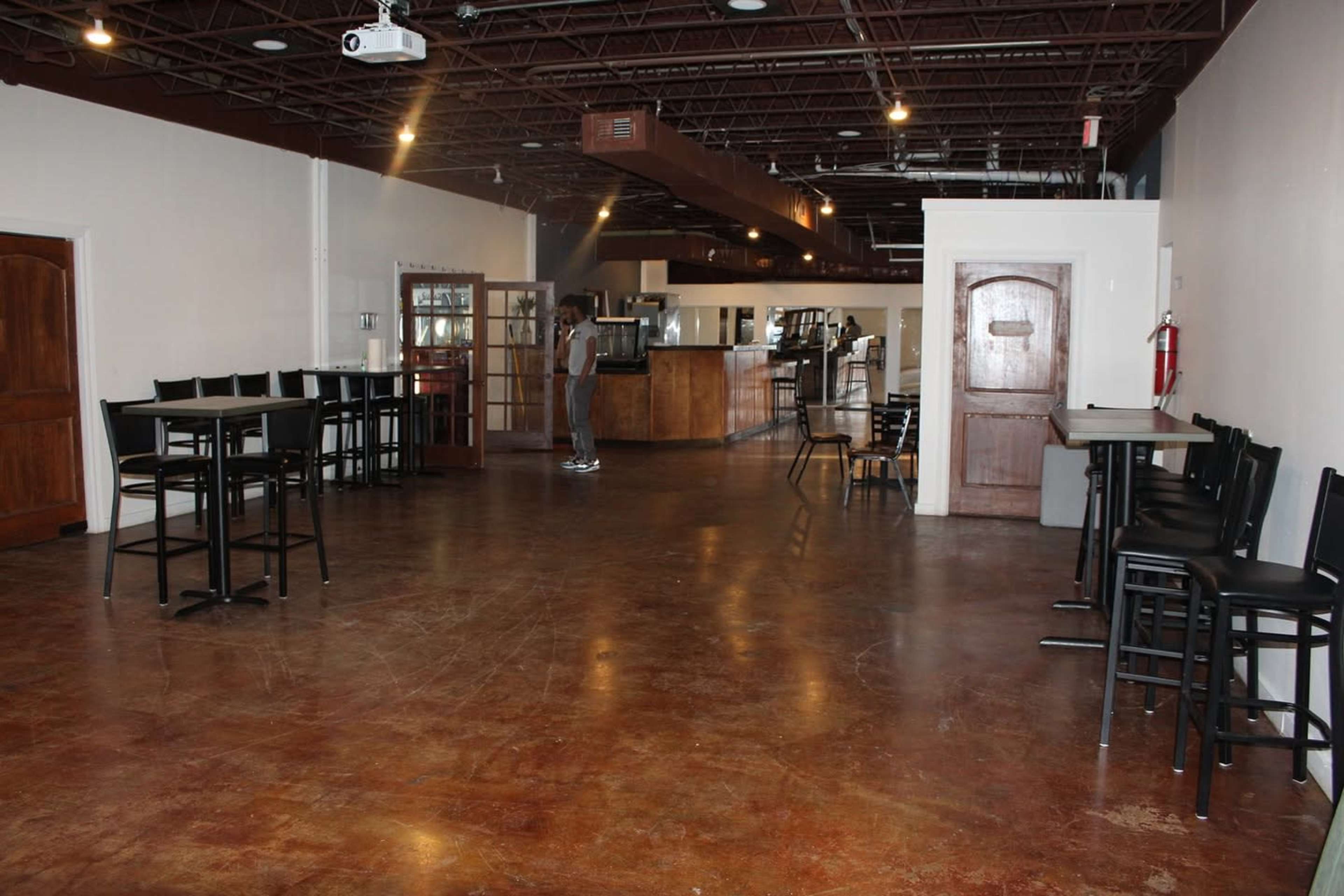 The image shows an empty dining area with several black chairs and tables, a bar counter in the background, and a person standing near the center.
