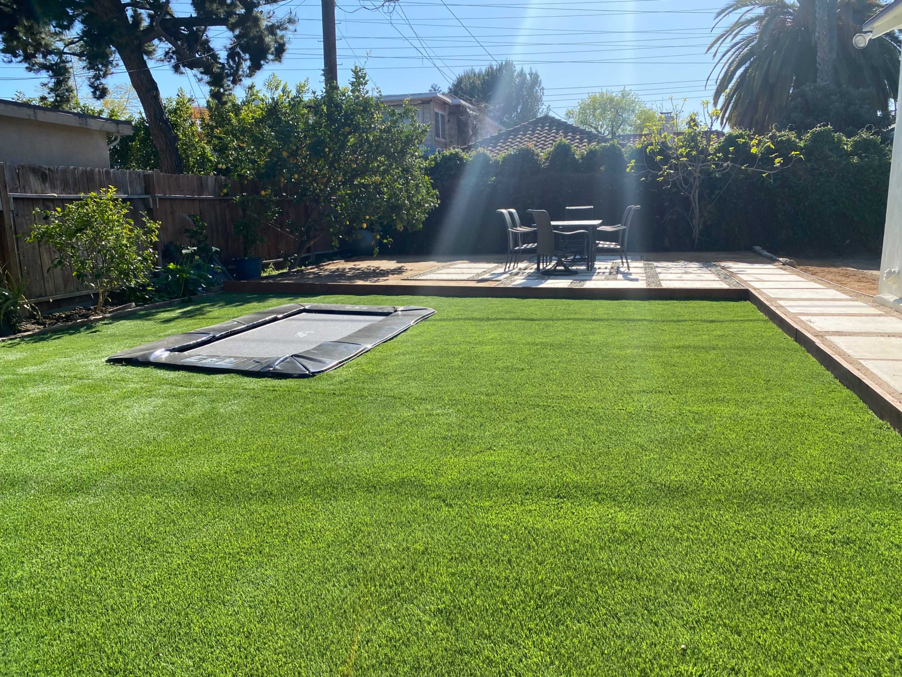 A backyard features a green lawn, a trampoline, and a patio set with chairs and a table.