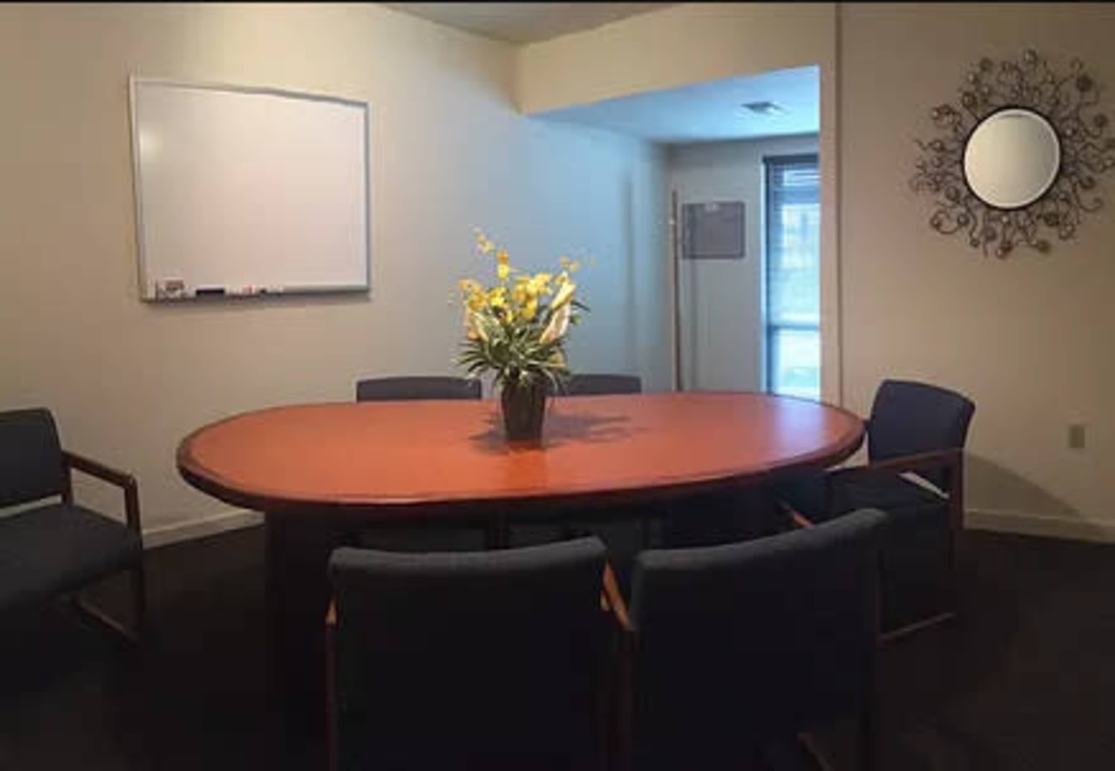 A conference room features a large oval table surrounded by four chairs, with a potted plant in the center and a whiteboard on the wall.