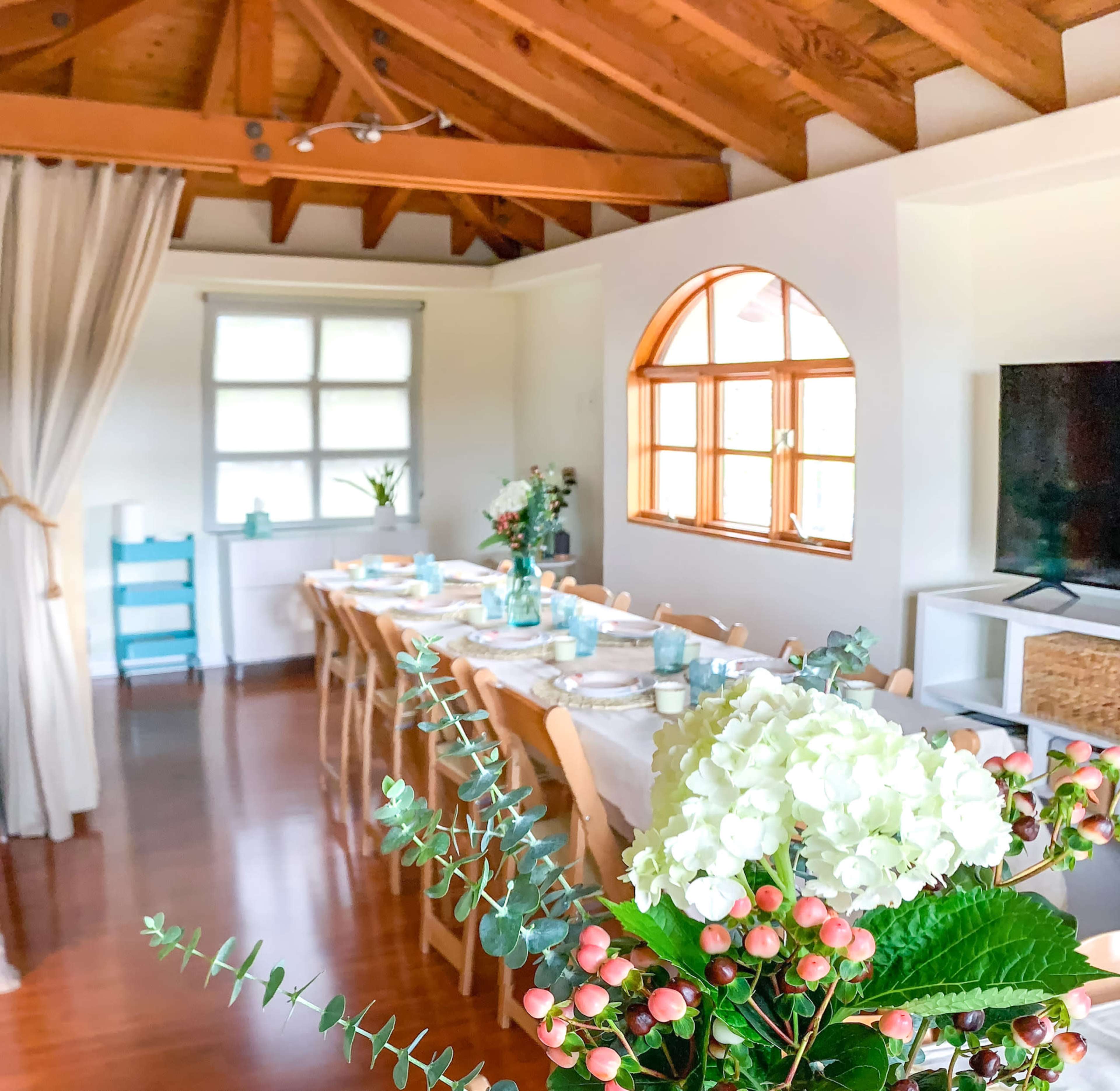A long dining table is set in a bright room with wooden beams and large windows, decorated with flowers and tableware.