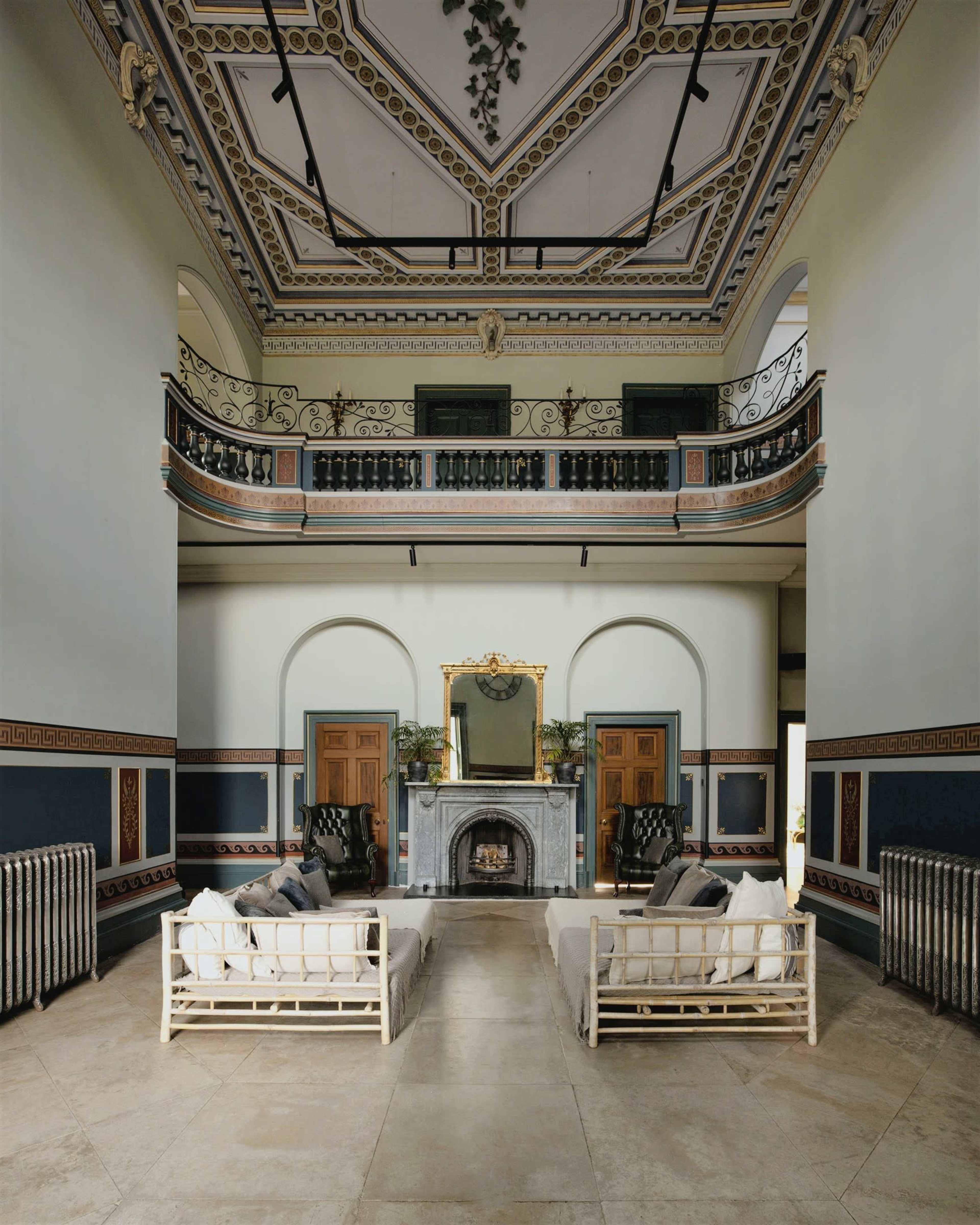 The image shows a spacious, grand living area featuring high ceilings, classical moldings, and two sets of white sofas arranged around a central fireplace.