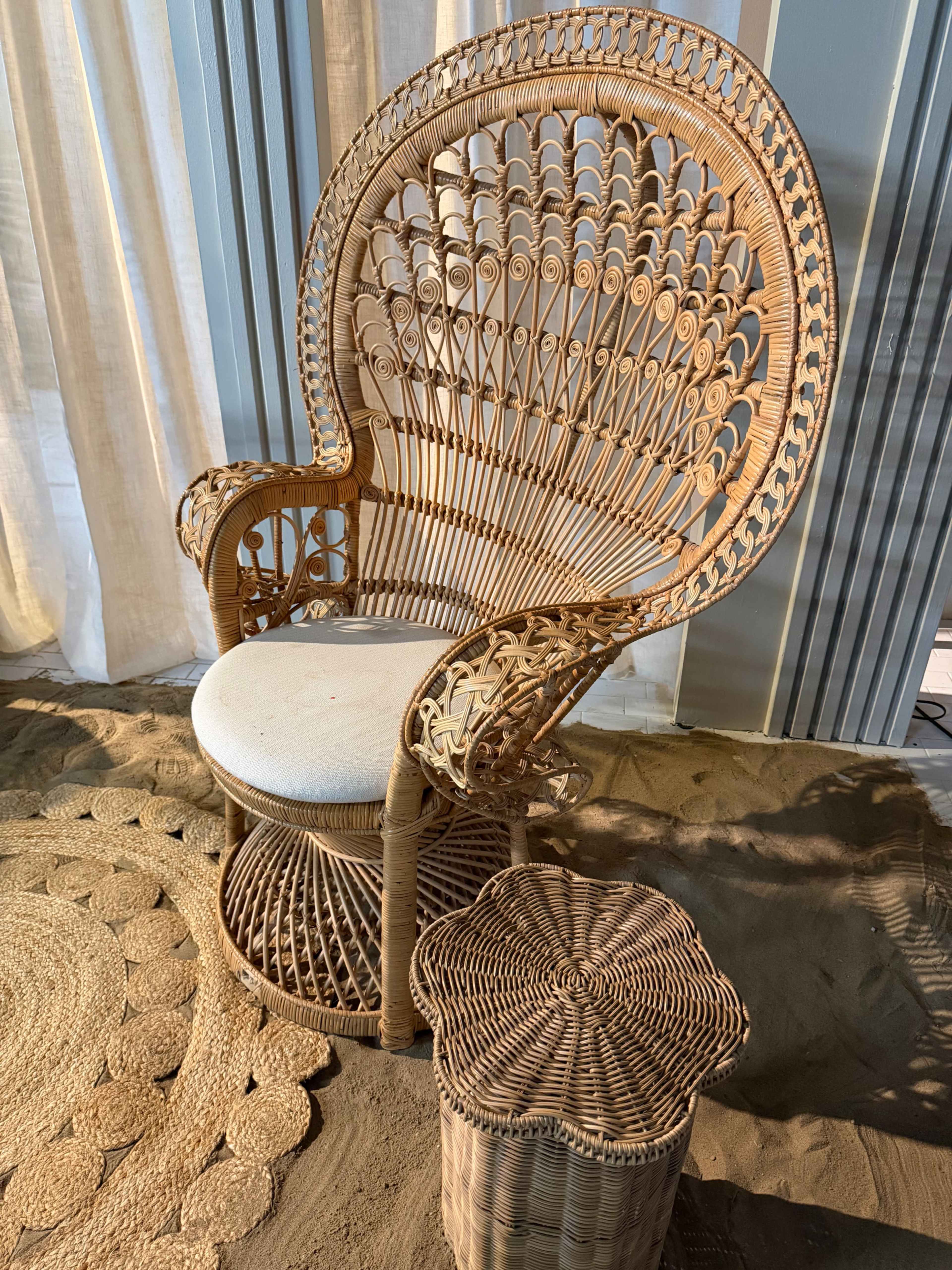 The image shows a large, intricately woven rattan chair with a round seat and a small matching side table beside it, set on a sandy surface with a textured circular mat underneath.