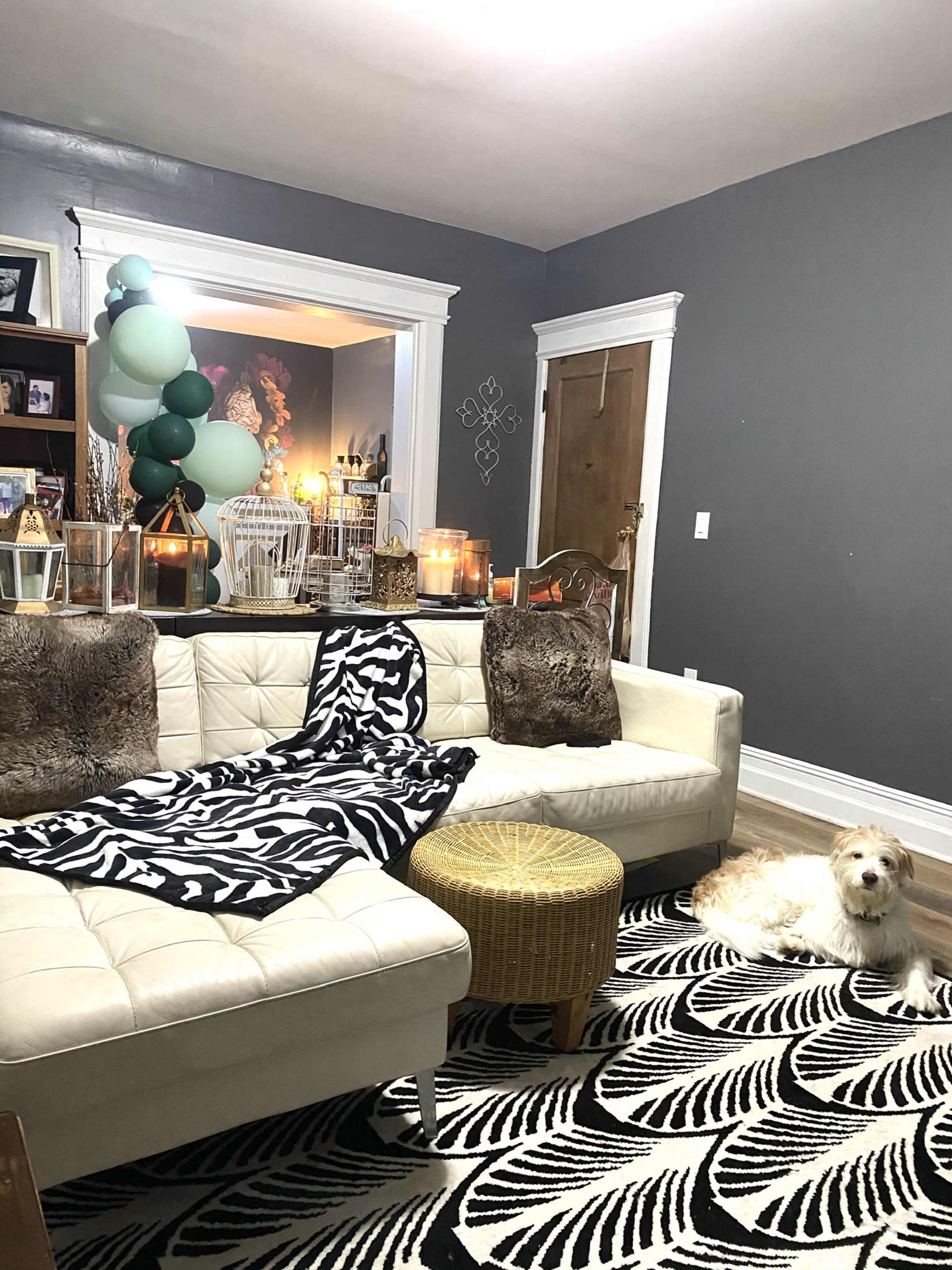 A cozy living room featuring a sectional sofa with a black and white blanket, a round ottoman, various decorative lanterns, and a small dog sitting on a patterned rug.