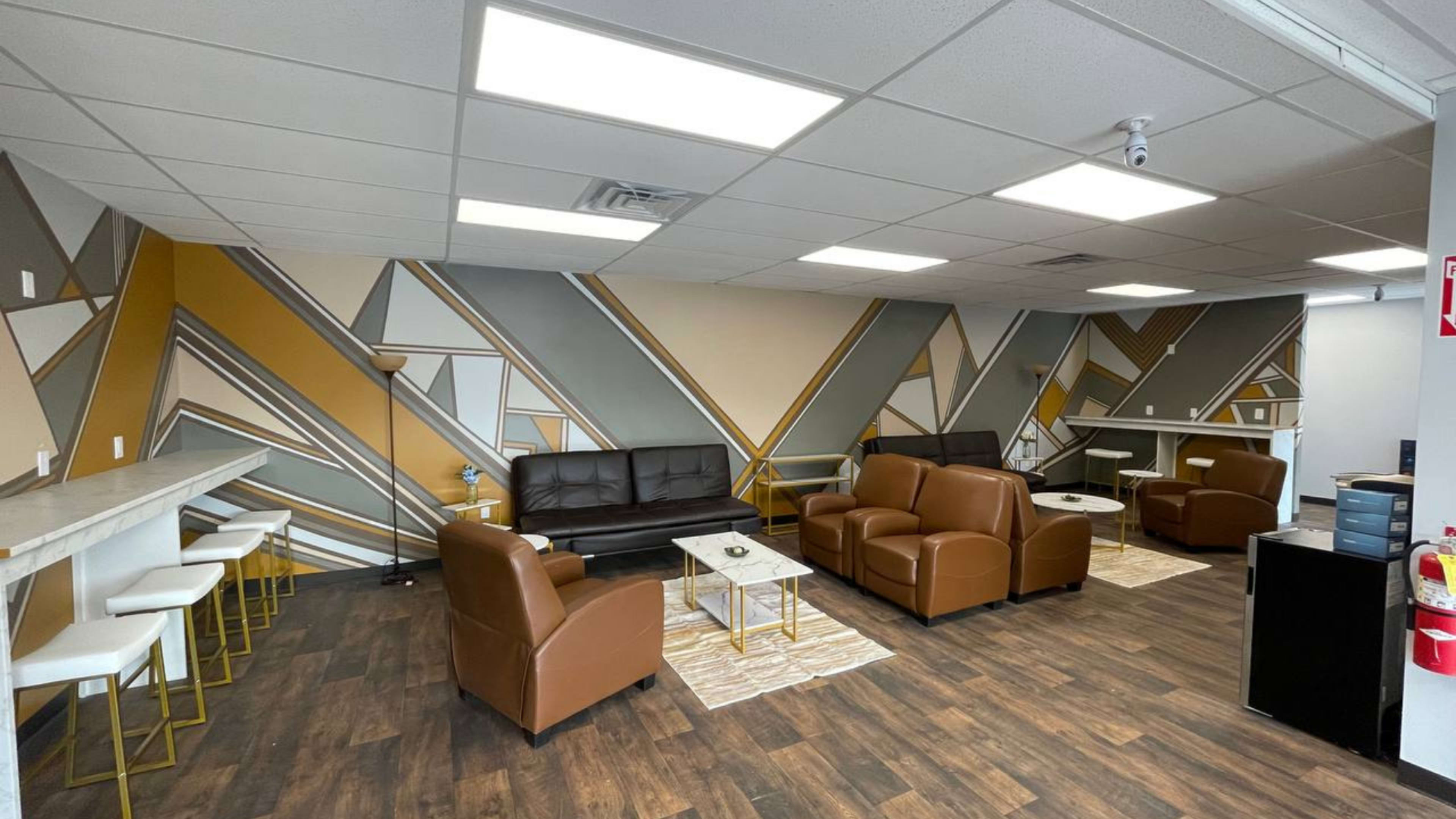 The image shows a modern lounge area featuring brown leather sofas, white side tables, and a geometric wall design in muted colors.