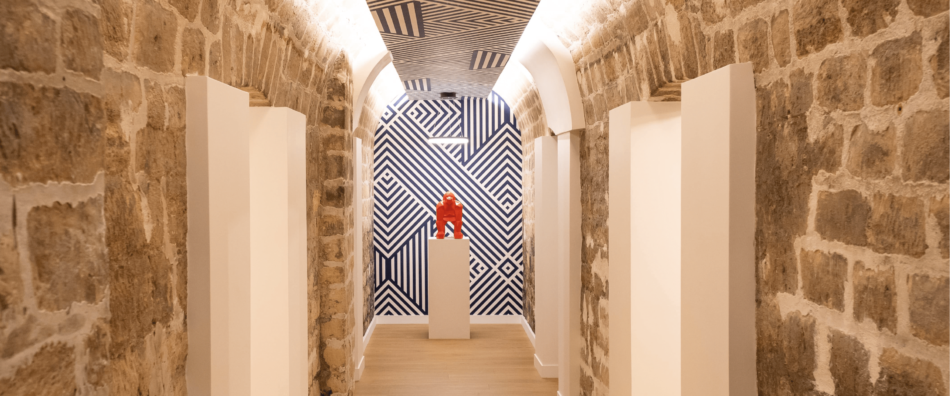 A narrow corridor with exposed stone walls, a geometric patterned ceiling, and a red sculpture positioned on a pedestal at the end.