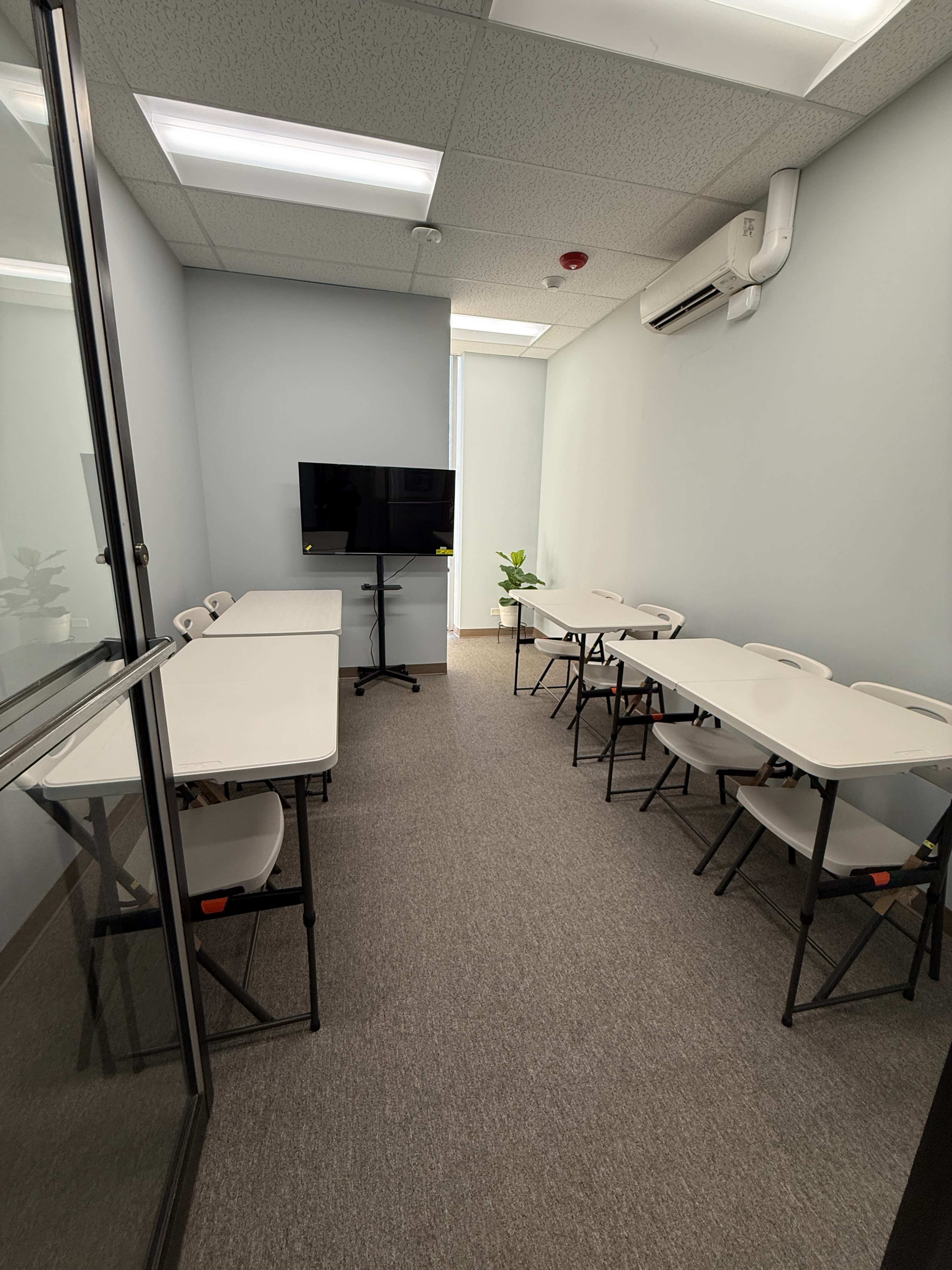 The image shows a small meeting room with several folding tables set up in rows, a television mounted on the wall, and a potted plant in the corner.