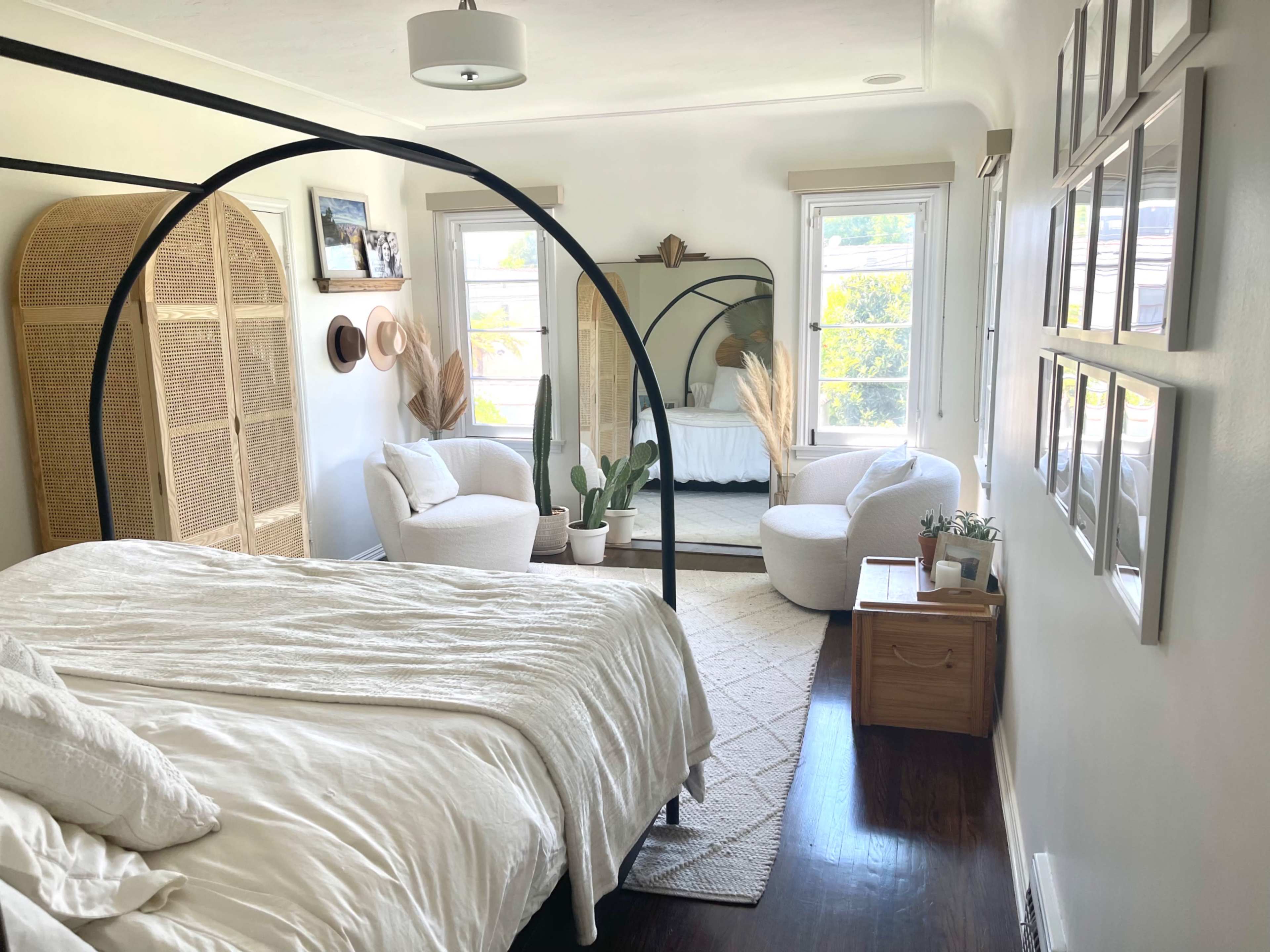The image shows a well-lit bedroom featuring a canopy bed, a cozy seating area with armchairs, and decorative plants arranged throughout the space.