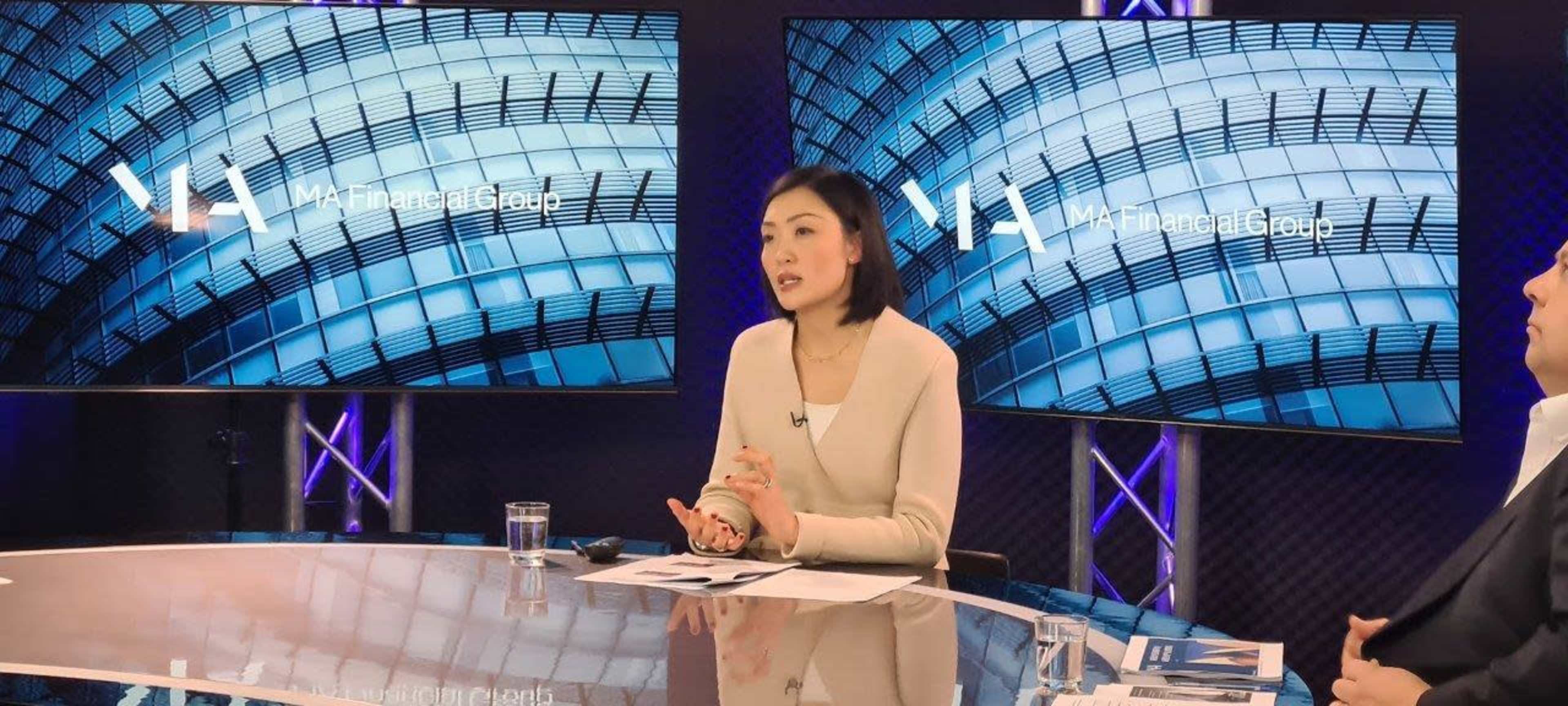 A woman is speaking at a desk with two display screens featuring a modern architectural design in the background.