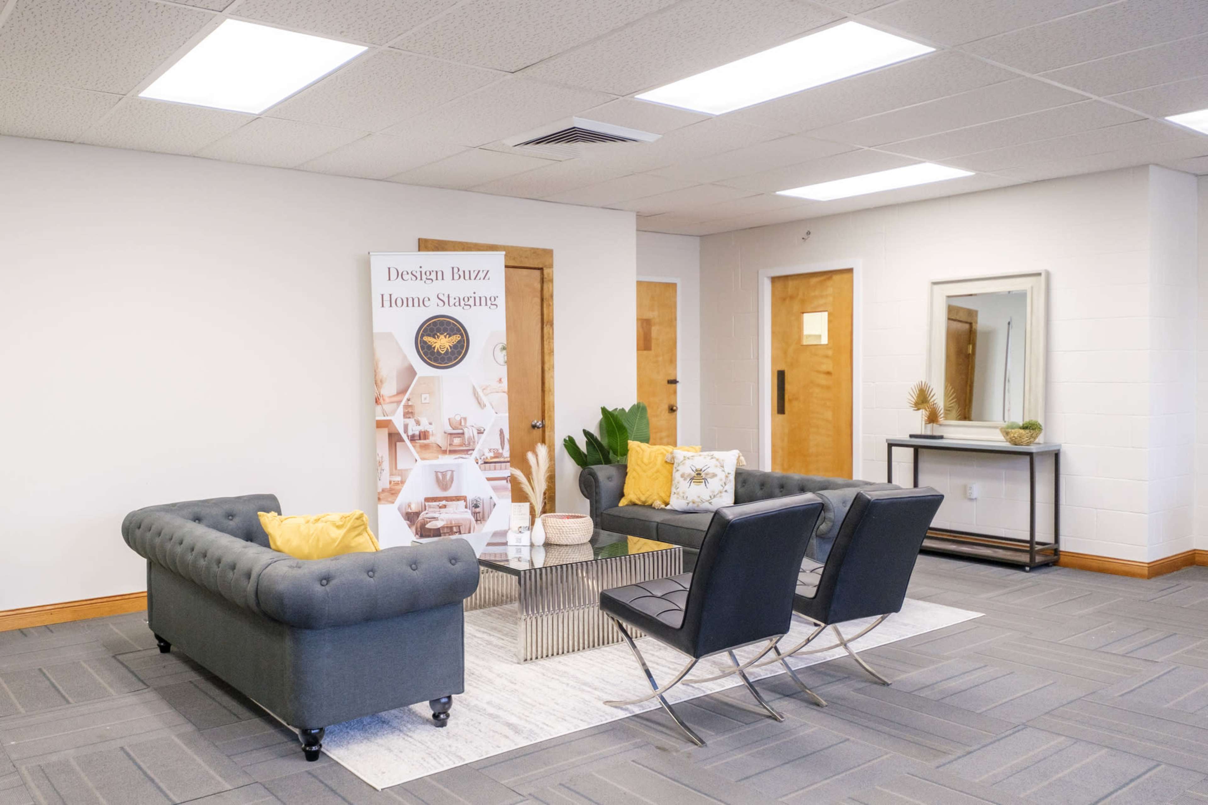 A modern waiting area with two gray couches, two black chairs, a coffee table in the center, and a banner advertising home staging against a light-colored wall.