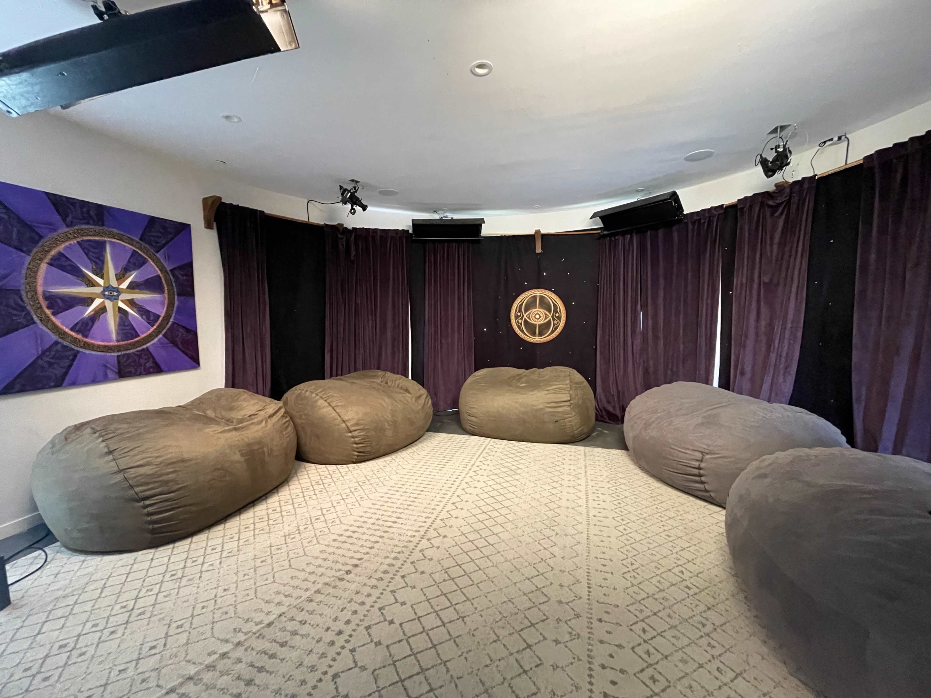 A circular room with dark curtains, large bean bags arranged around a circular rug, and a decorative wall hanging featuring a symbol.