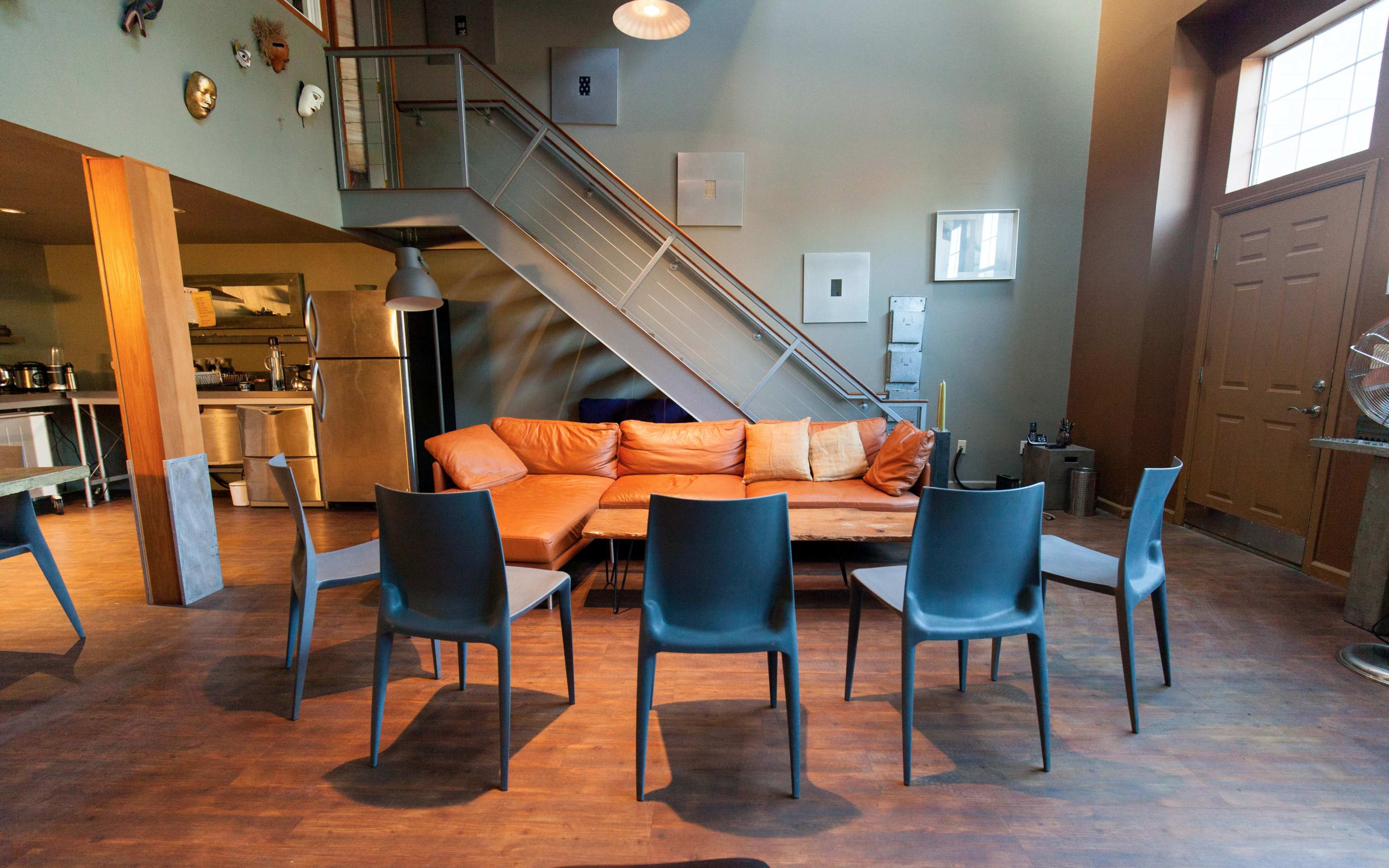A modern living space with a staircase in the background, an orange sectional sofa, and several gray dining chairs arranged around a wooden table.