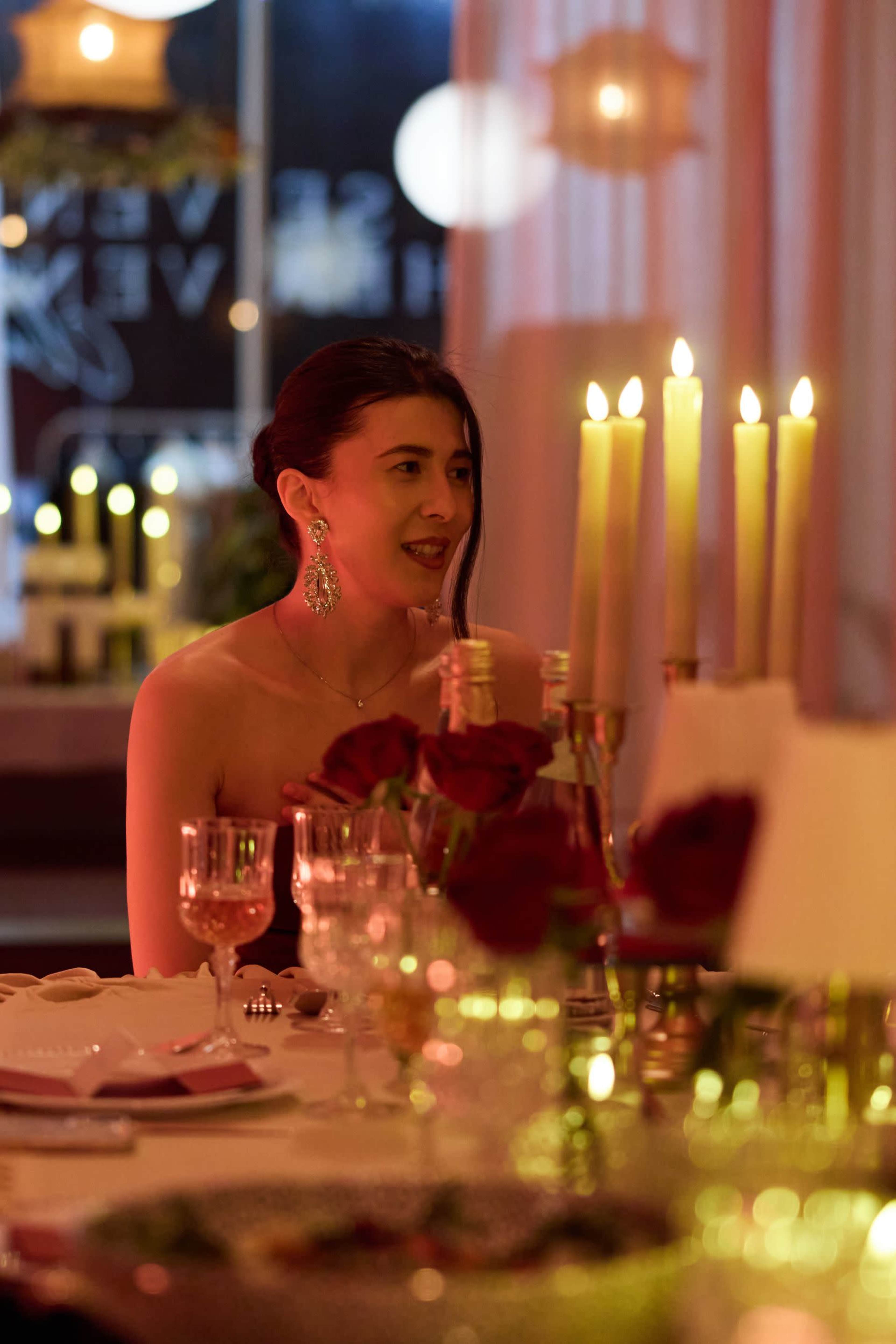 A woman sits at a elegantly set dining table with lit candles and roses in a warm, softly illuminated setting.