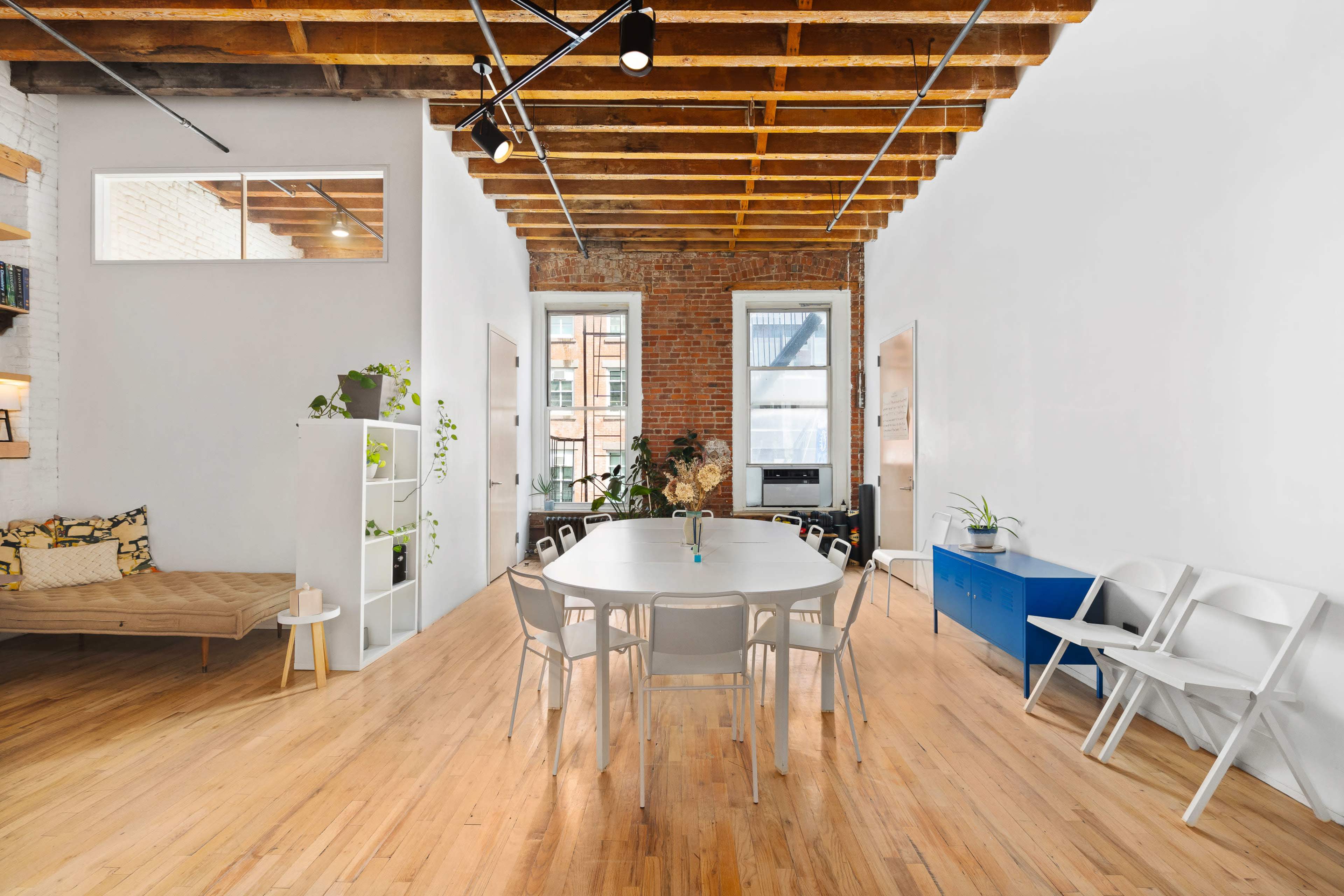 The image shows a modern, open-concept communal space featuring a round table surrounded by chairs, wooden beams on the ceiling, and large windows displaying brick walls.