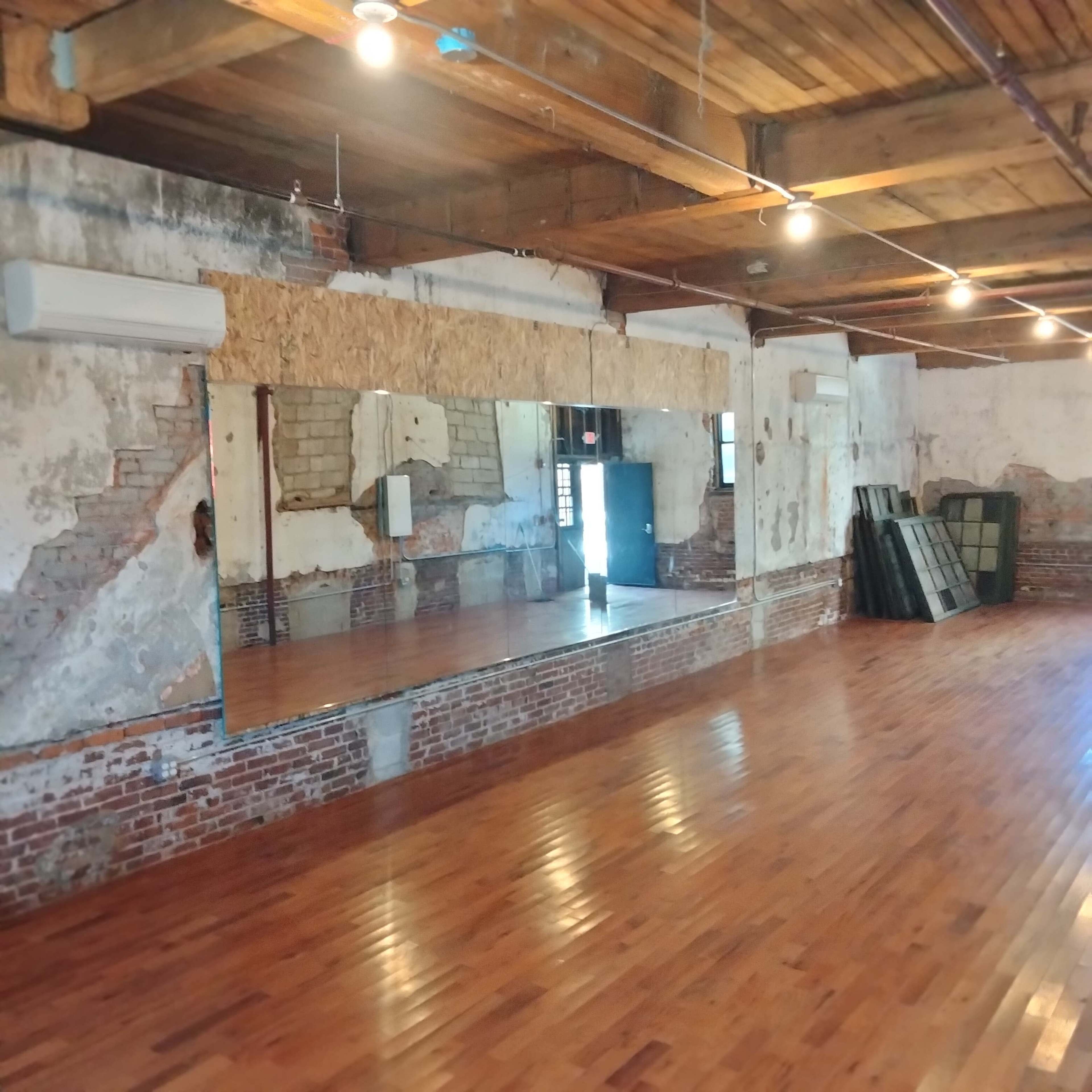 The image shows a spacious room with wooden floors, exposed brick walls, and a large mirror mounted on a wooden frame.