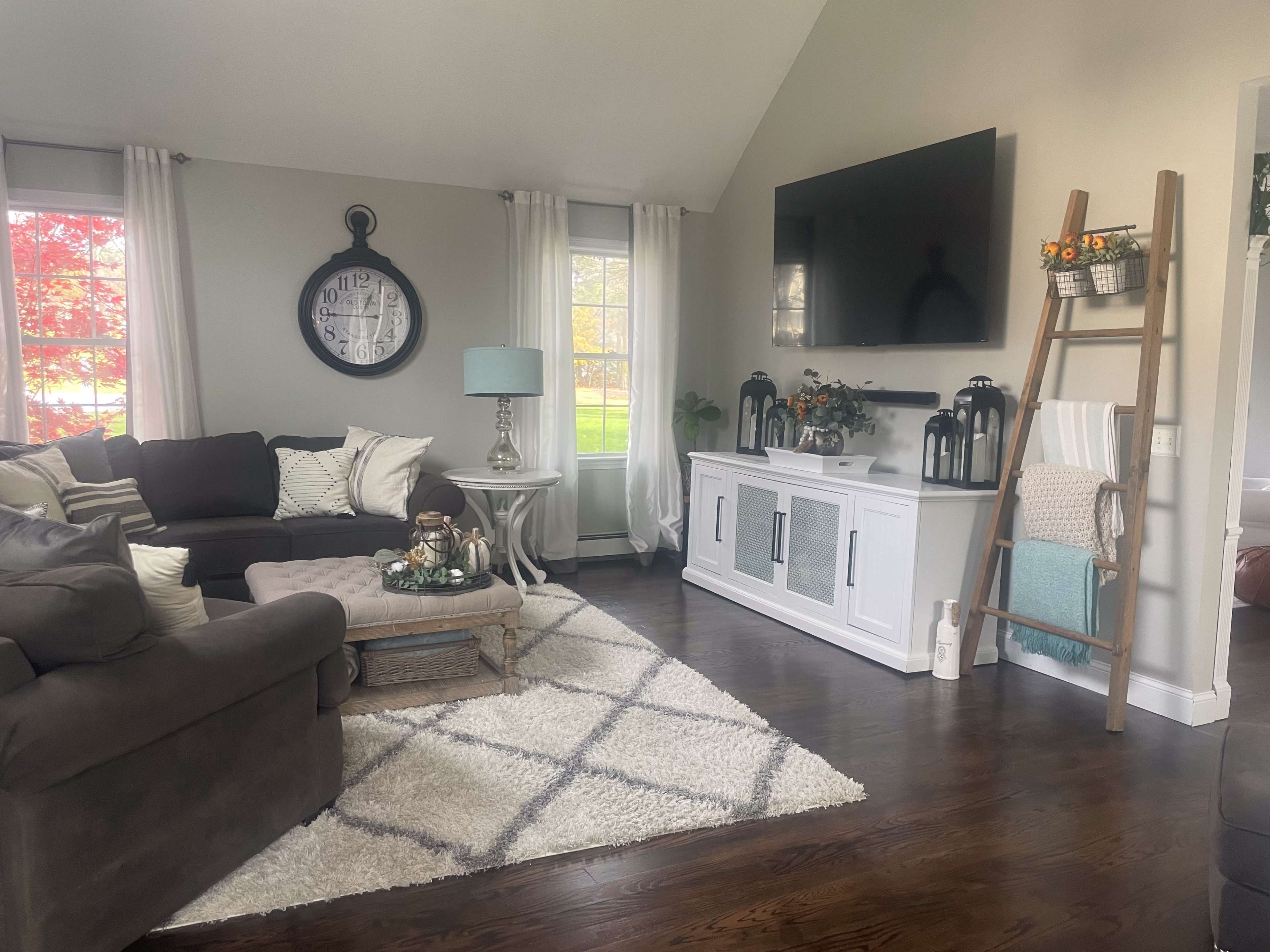 The living room features a sectional sofa, a coffee table on a patterned rug, a wall-mounted TV, and decorative elements like lanterns and a ladder shelf.