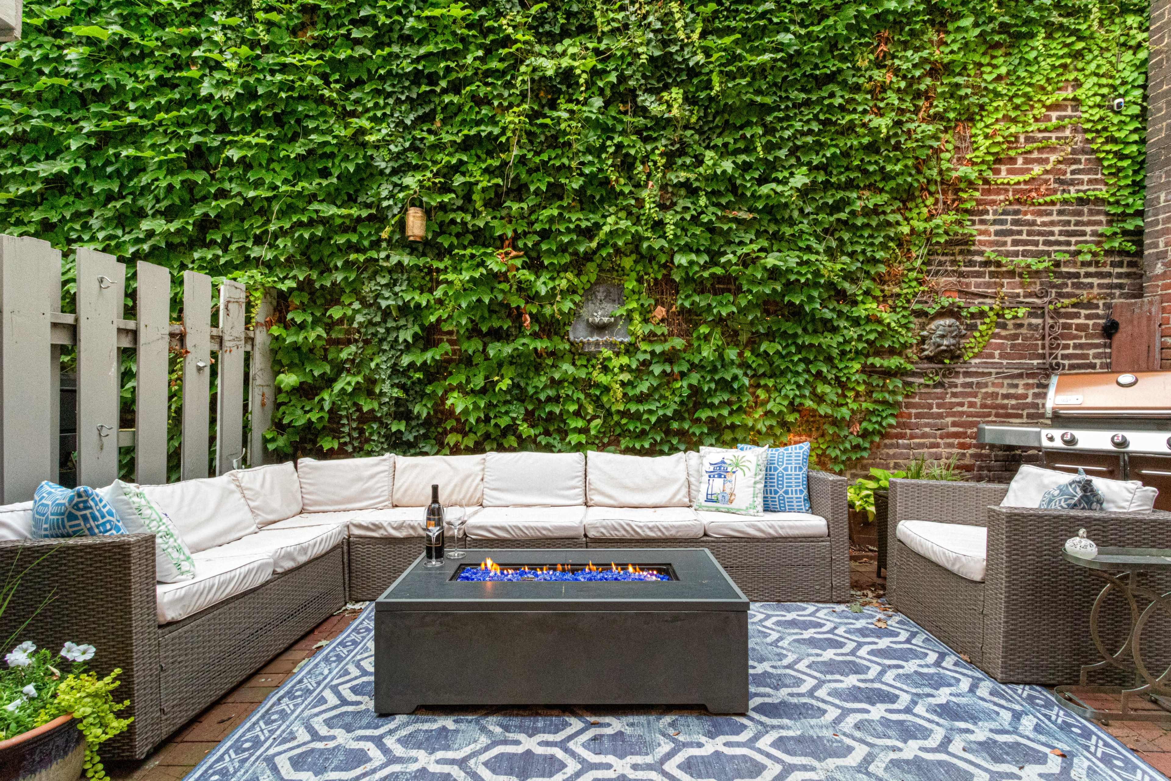 A cozy outdoor seating area features a sectional sofa facing a fire table, surrounded by lush green ivy-covered walls and a nearby barbecue grill.
