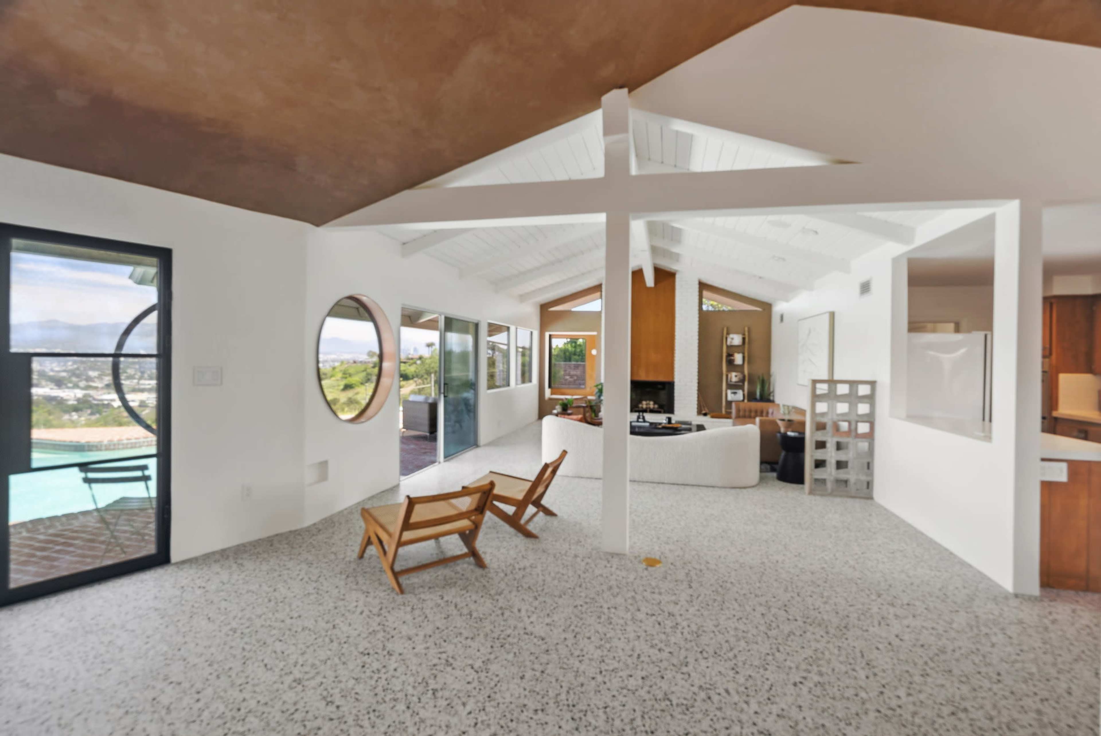 The image shows a modern, open-concept living space featuring a light-colored floor, large windows, and a pool visible outside.