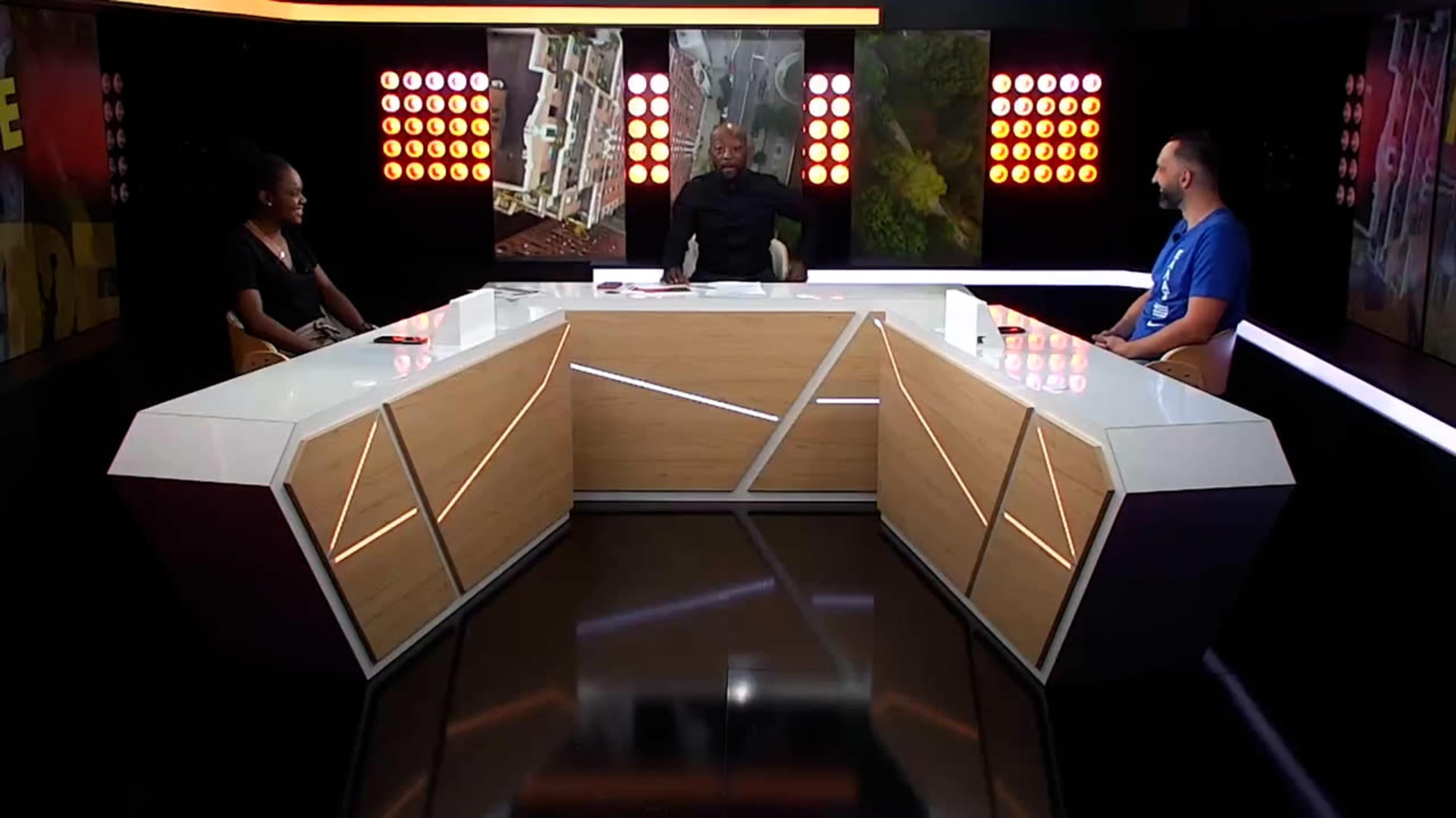 A television studio features three people sitting at a modern, angular desk surrounded by large screens displaying images.