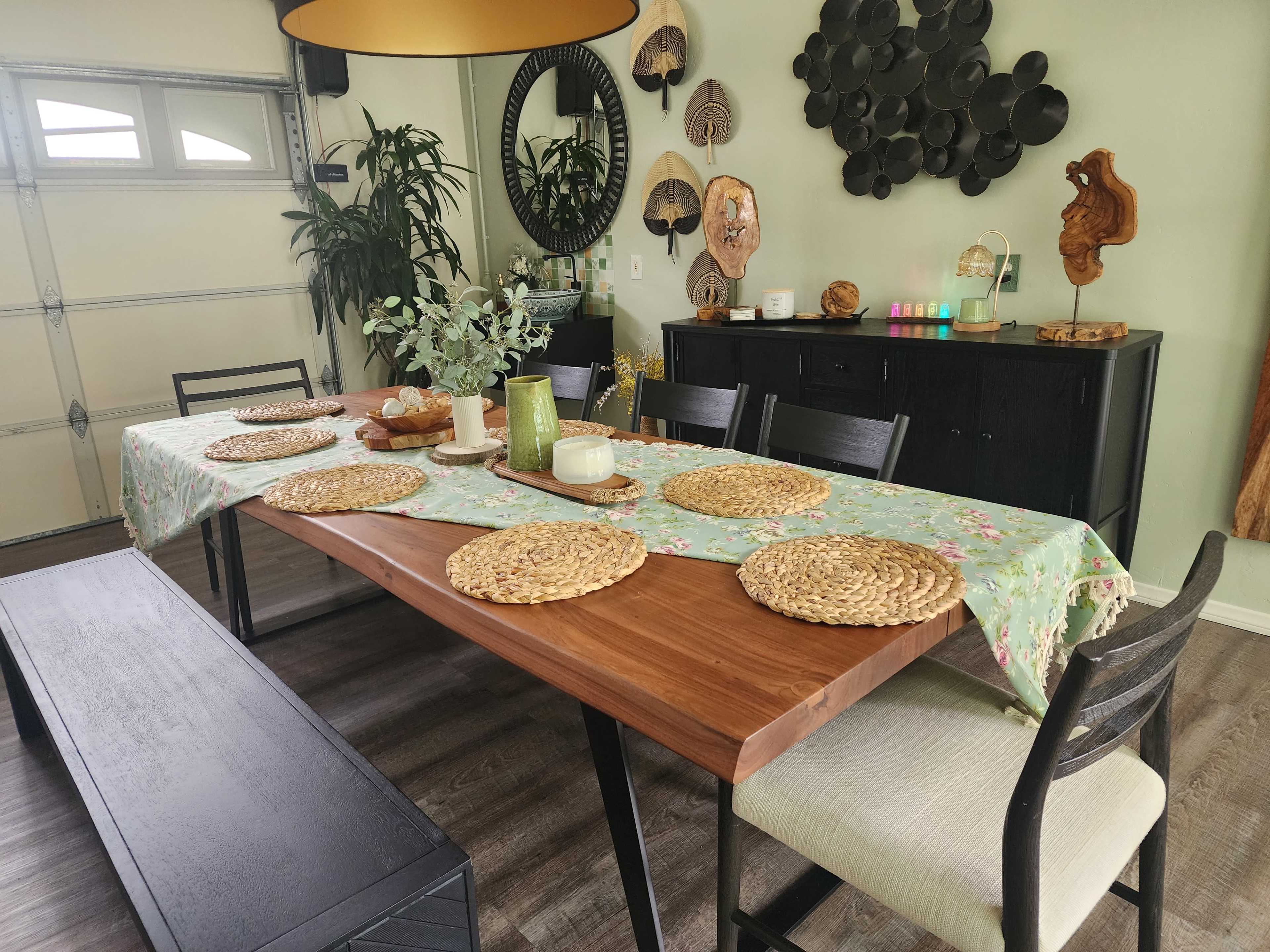 A dining table is set with woven placemats and a floral tablecloth in a well-decorated room featuring plants and wall decor.