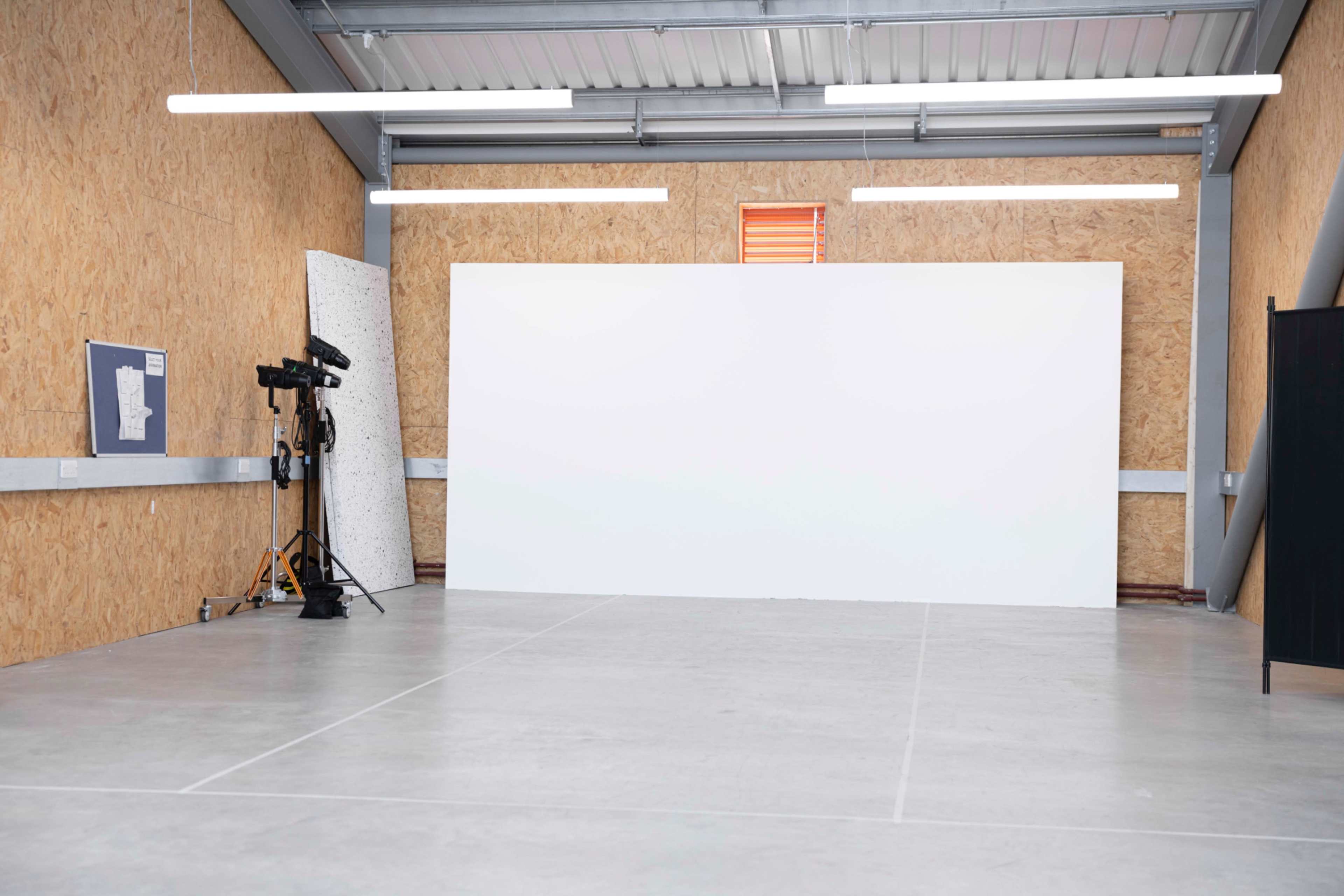 The image shows an empty studio space with a large white wall, concrete flooring, and wooden walls.