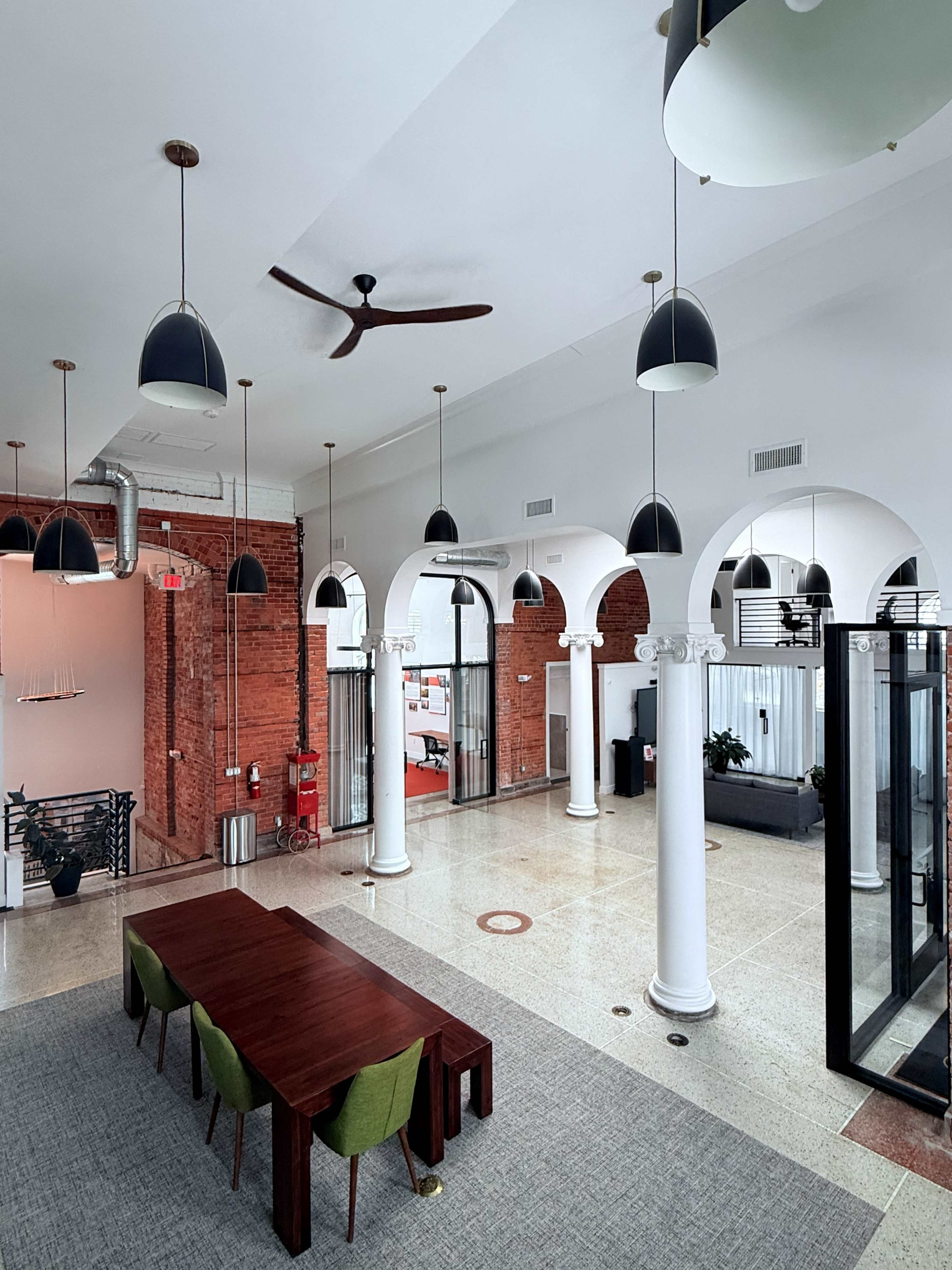 The image shows a spacious office interior with a large wooden table, green chairs, tall white columns, and modern lighting fixtures hanging from the ceiling.
