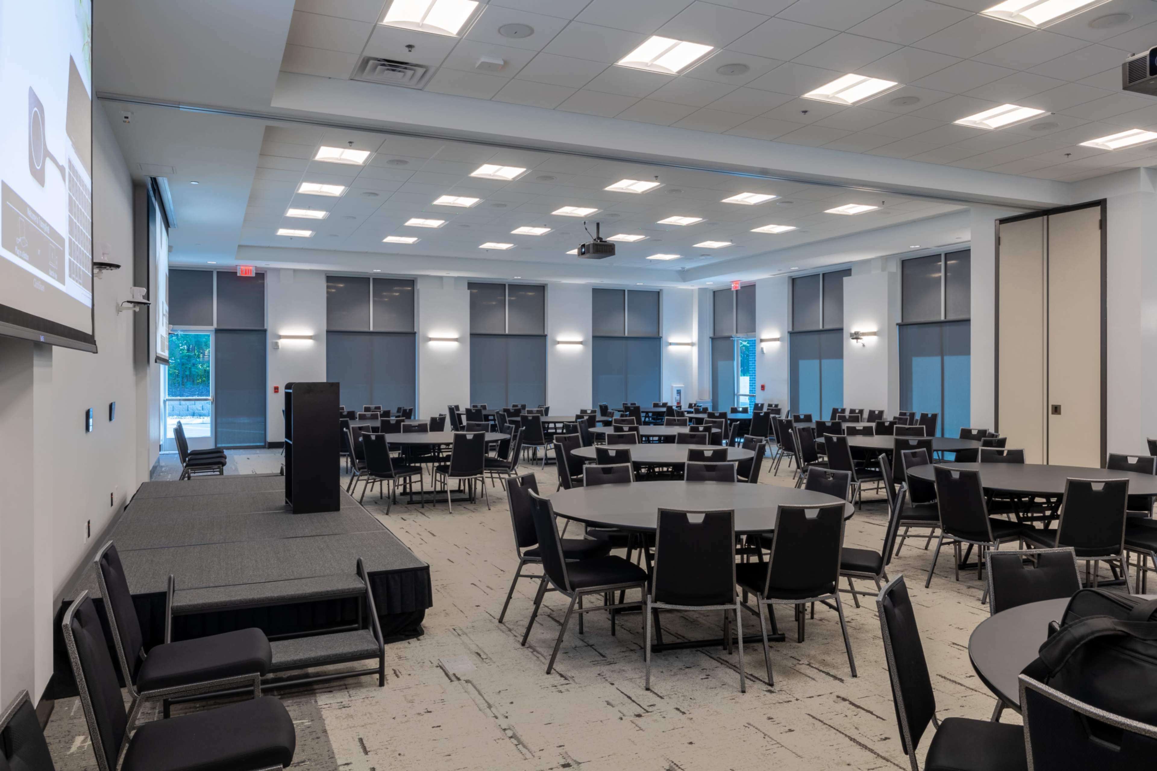 The image shows a spacious conference room with multiple round tables set up, a podium at the front, and large windows with shades.